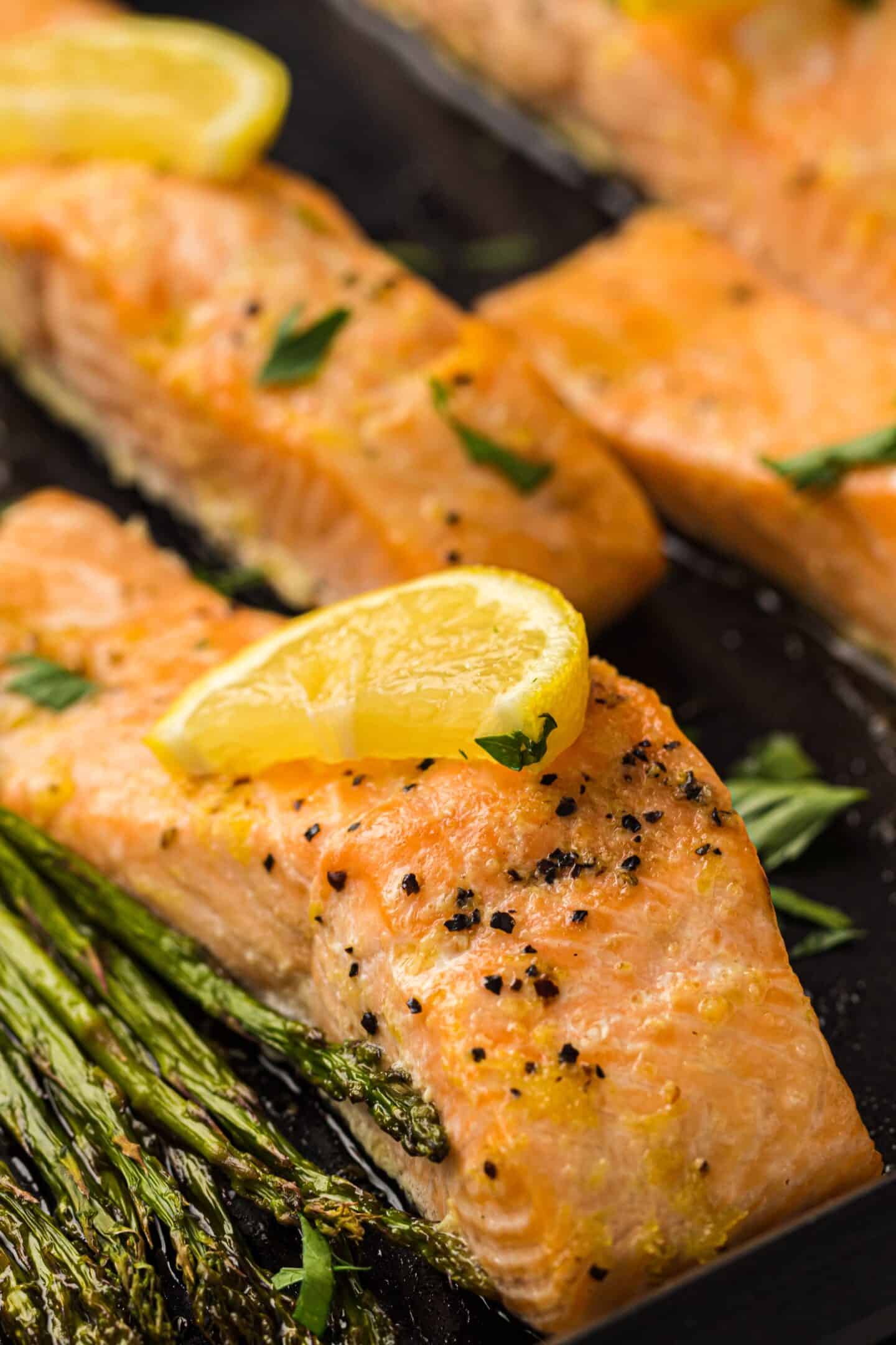 Close-up of baked salmon fillets topped with lemon wedges and black pepper, served with roasted asparagus and golden roasted potatoes on a dark tray.