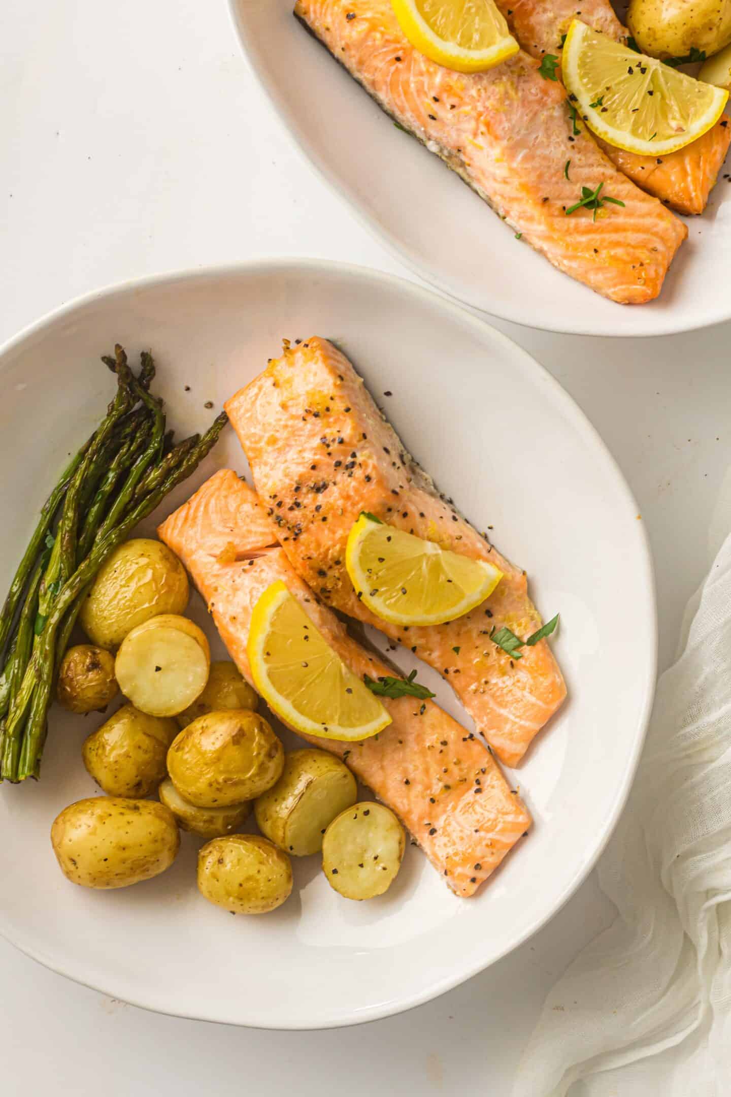 Two white plates with Sheet Pan Salmon fillets topped with lemon slices, served alongside roasted potatoes and asparagus spears on a white surface.