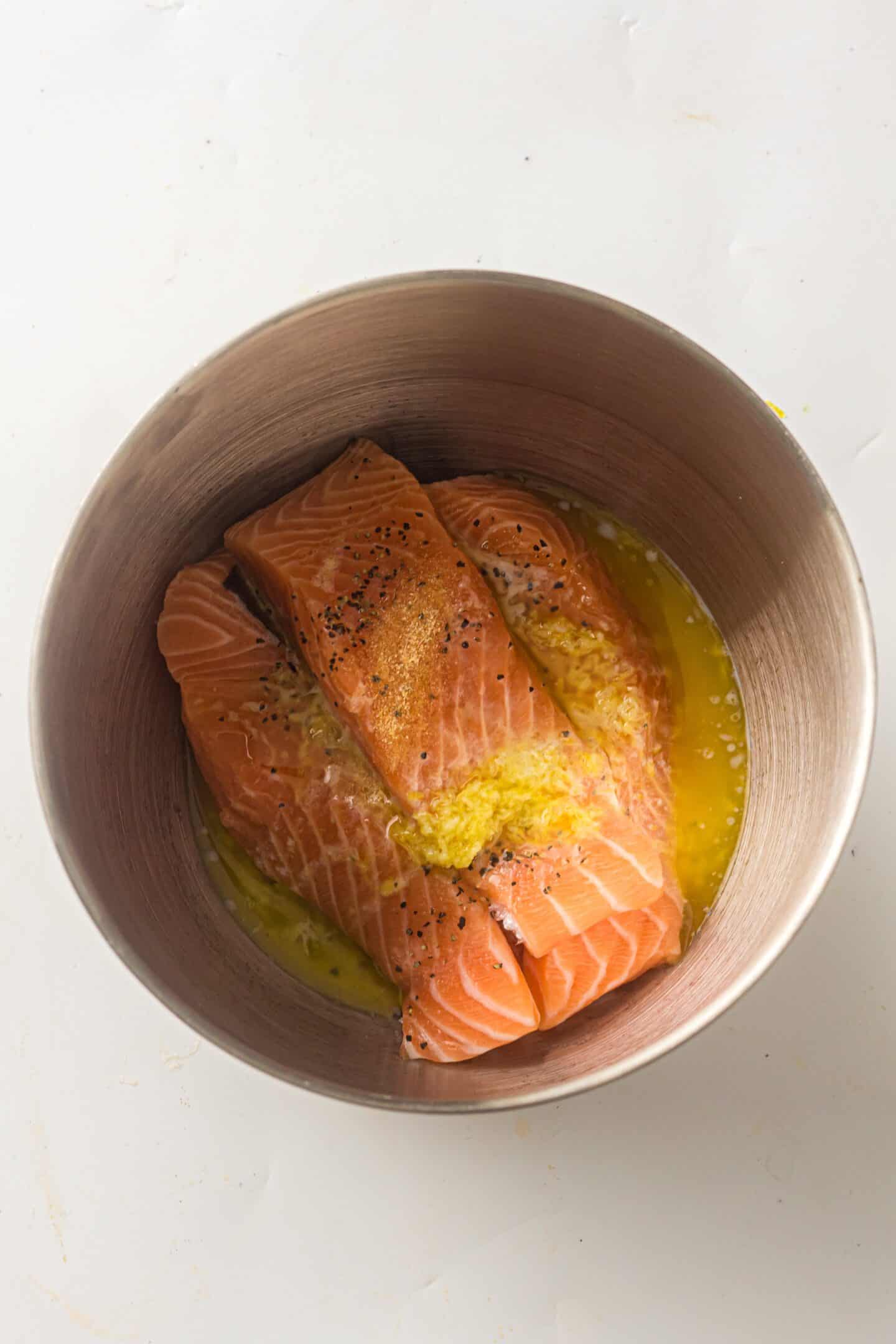 Three raw salmon fillets in a metal bowl with melted butter, minced garlic, lemon zest, and black pepper on top—perfect for making Sheet Pan Salmon with roasted potatoes and asparagus.