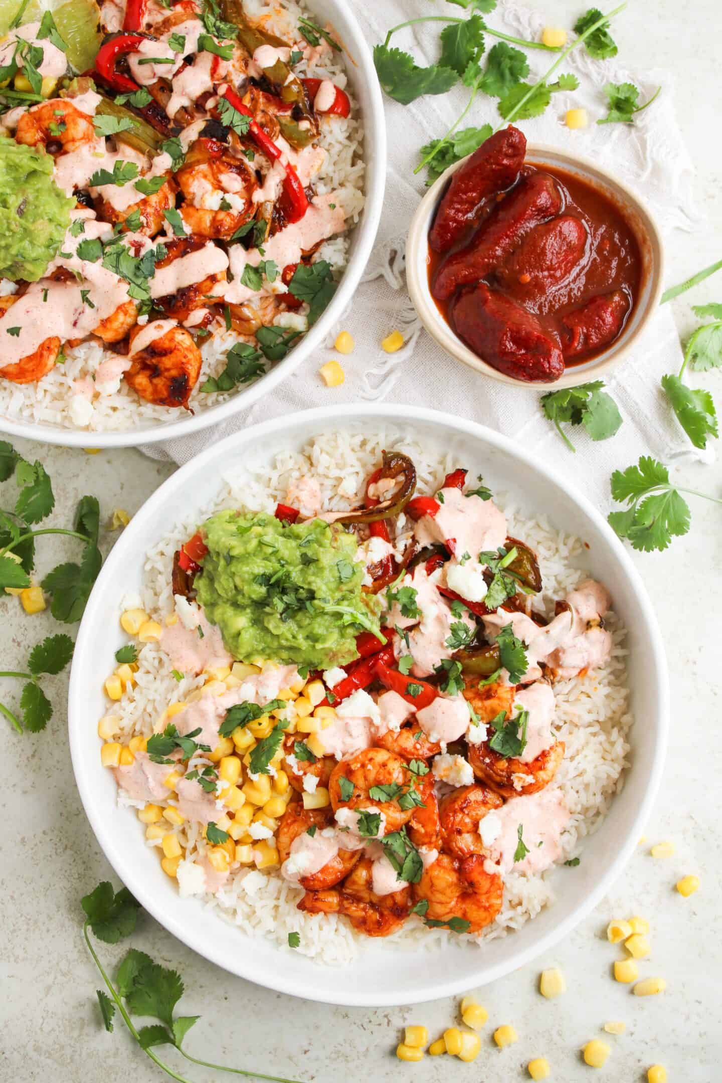 Two Shrimp Fajita Bowls topped with shrimp, corn, sautéed peppers, guacamole, and chipotle sauce, served alongside a dish of sauced peppers and scattered cilantro and corn kernels.