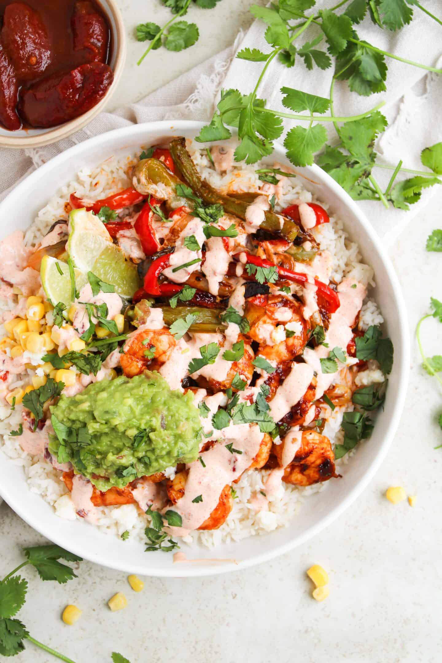 A bowl of rice topped with grilled vegetables, chicken, guacamole, corn, pink sauce, lime wedges, and fresh cilantro—served with a small bowl of chipotle sauce on the side for a flavorful fajita recipe twist.