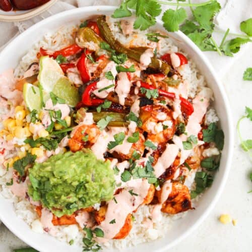 A bowl of rice topped with grilled vegetables, chicken, guacamole, corn, pink sauce, lime wedges, and fresh cilantro—served with a small bowl of chipotle sauce on the side for a flavorful fajita recipe twist.