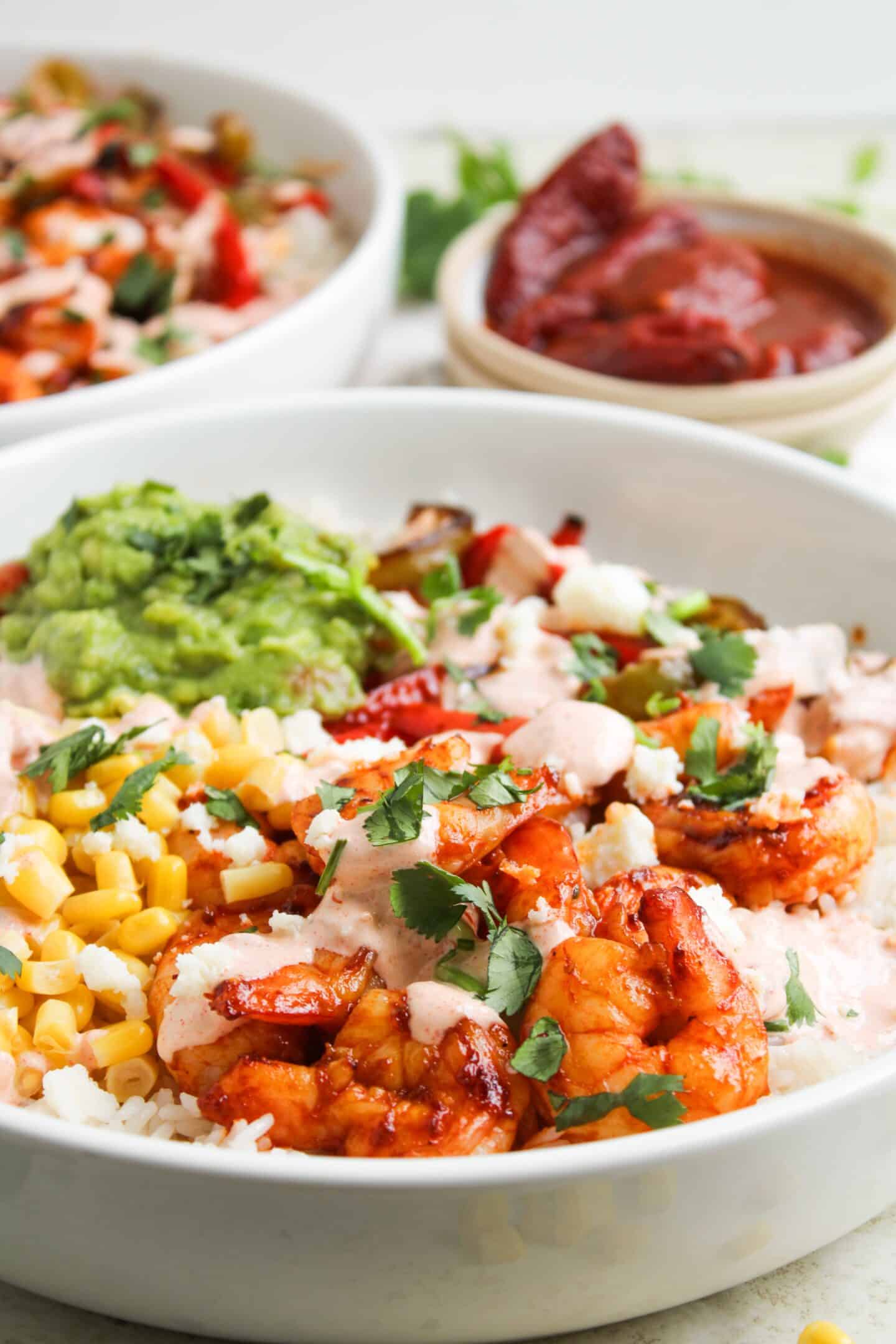 A bowl of rice topped with grilled shrimp, corn, guacamole, chopped herbs, and creamy chipotle sauce, with a bowl of salsa and another dish in the background—perfect for fans of Shrimp Fajita Bowls.