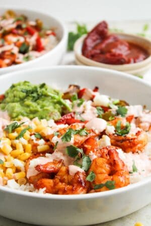 A bowl of rice topped with grilled shrimp, corn, guacamole, chopped herbs, and creamy chipotle sauce, with a bowl of salsa and another dish in the background—perfect for fans of Shrimp Fajita Bowls.