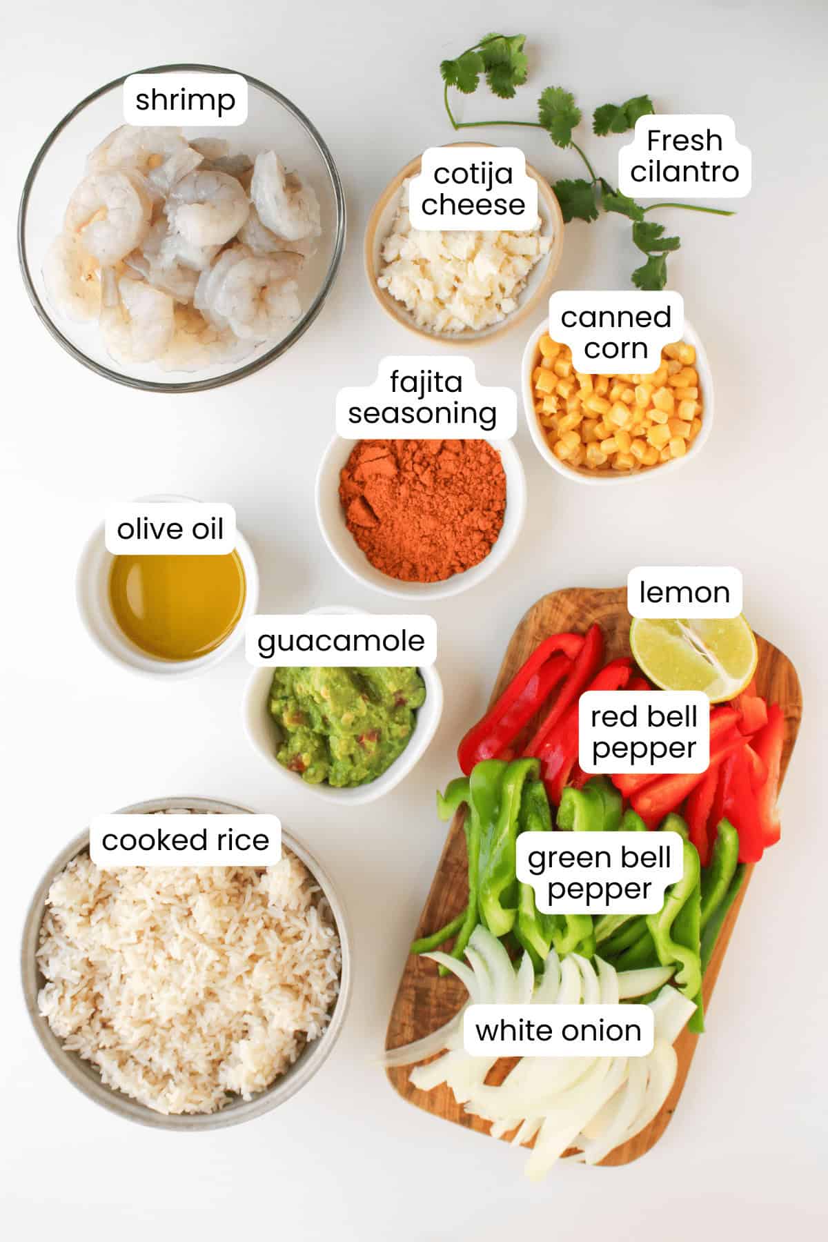Overhead view of Shrimp Fajita Bowls with cotija cheese, fresh cilantro, canned corn, olive oil, fajita seasoning, guacamole, lemon, bell peppers, white onion, cooked rice, and a drizzle of Chipotle Sauce.