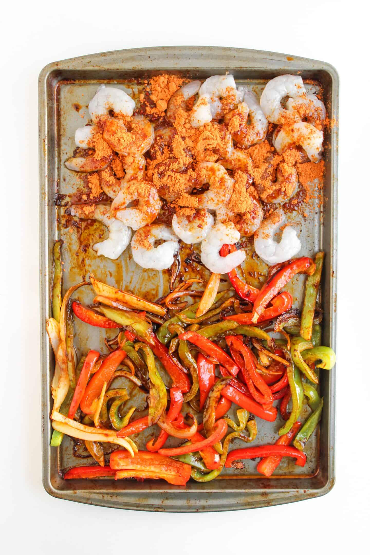 A baking sheet with raw shrimp topped with seasoning on one half and cooked red and green bell pepper strips on the other—perfect for assembling Shrimp Fajita Bowls or serving with a tangy chipotle sauce.