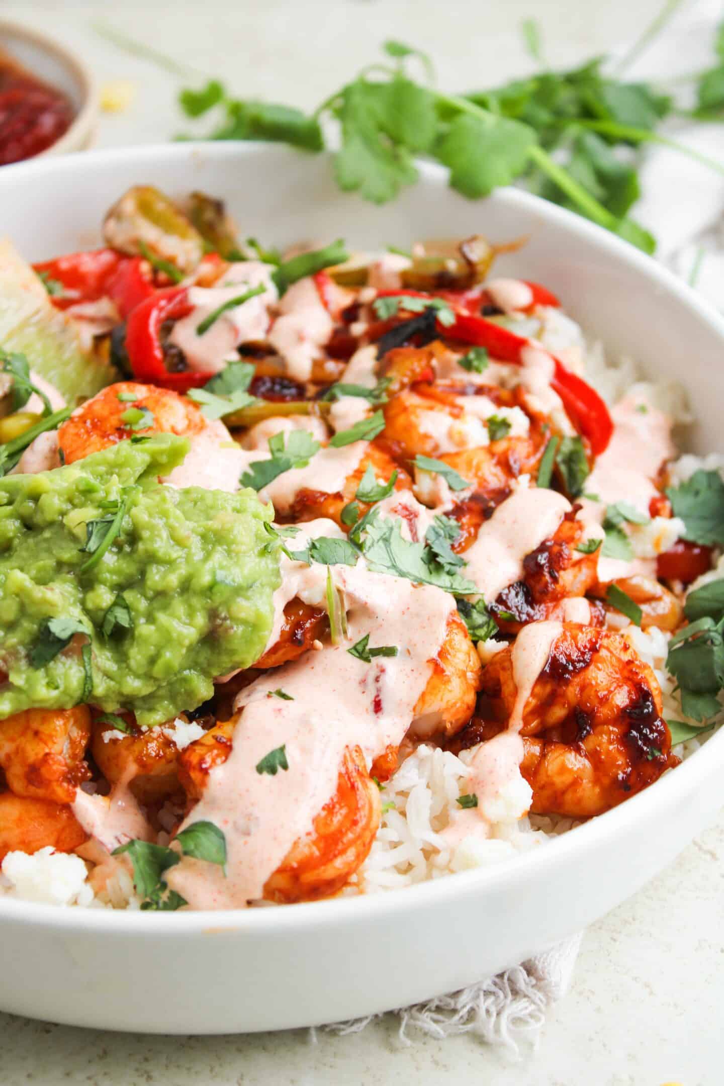 A bowl of white rice topped with shrimp, sautéed peppers, creamy chipotle sauce, guacamole, and chopped cilantro—perfect for anyone craving delicious Shrimp Fajita Bowls.