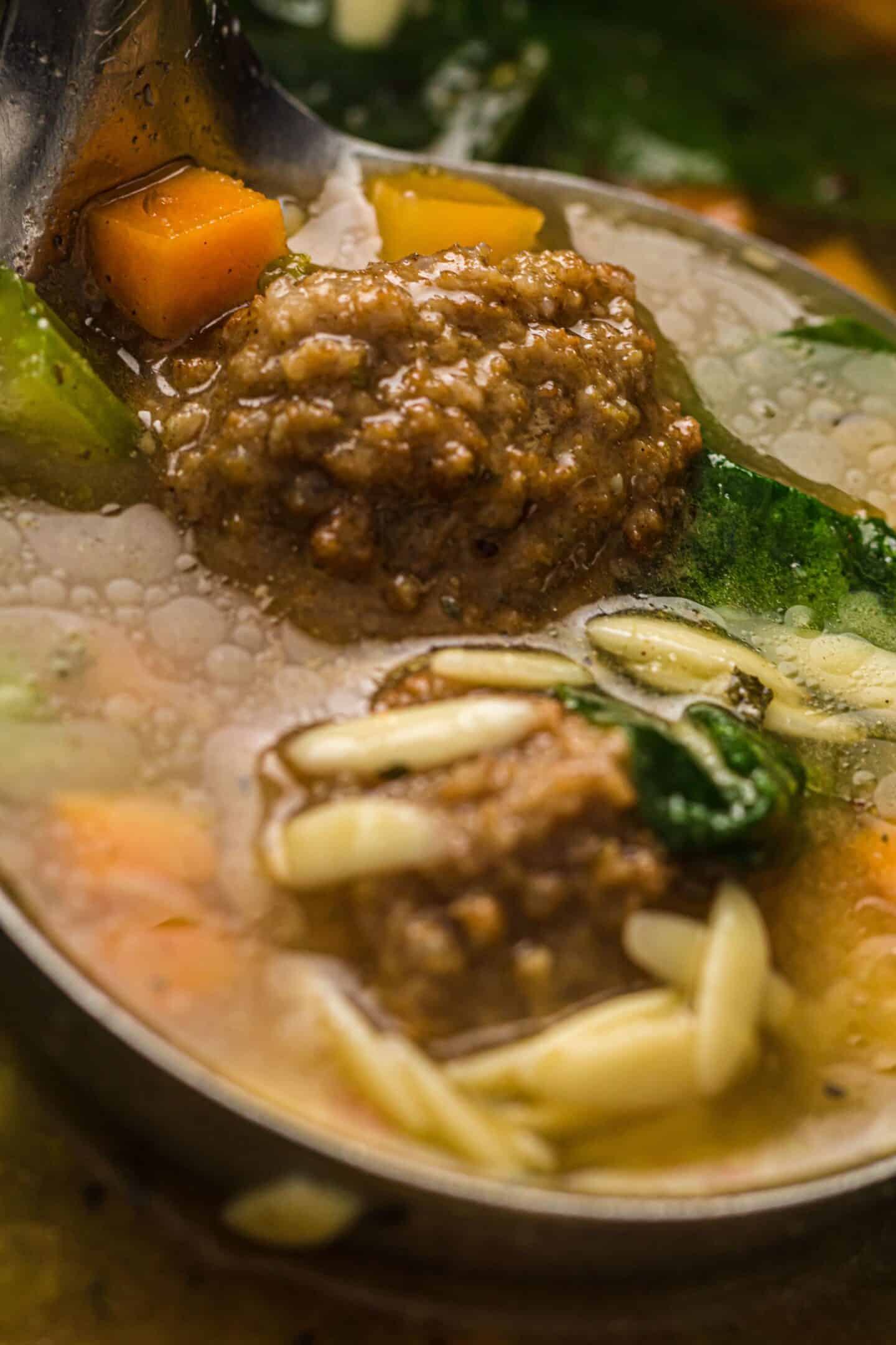 A close-up of a ladle holding meatballs, vegetables, greens, and orzo pasta in a clear broth—classic Italian Wedding Soup made easy as a one-pot meal.