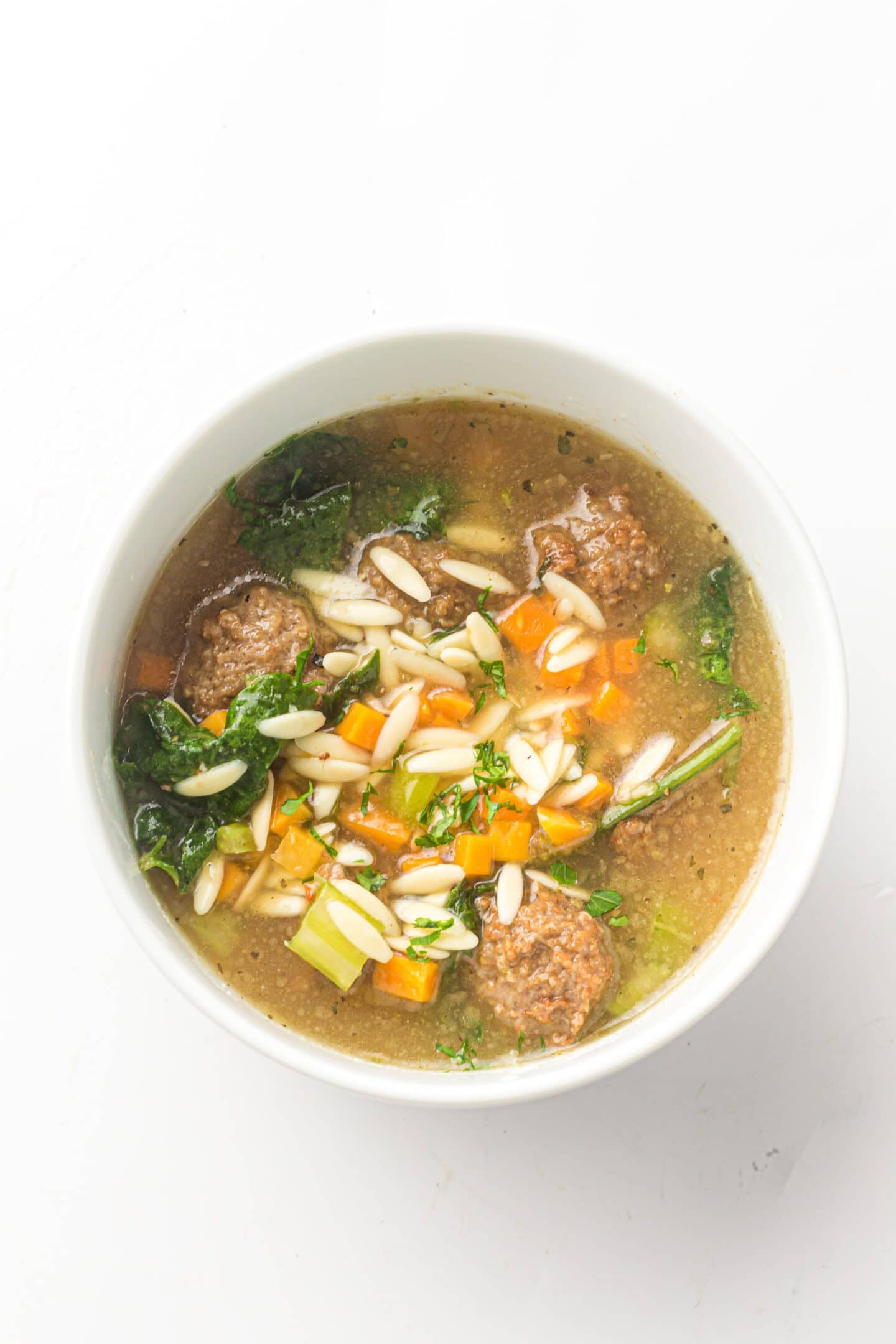 A bowl of Italian Wedding Soup containing meatballs, orzo pasta, sliced carrots, celery, kale, and broth is photographed from above on a white background.