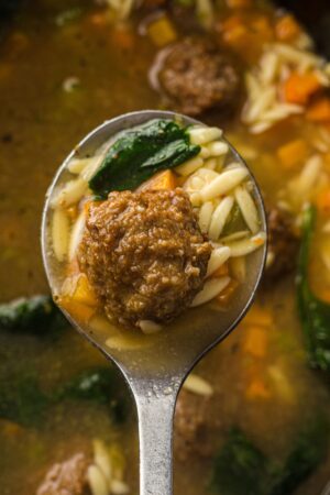 A close-up of a spoon holding a meatball, orzo pasta, spinach, and vegetables in broth above a bowl of Italian Wedding Soup.