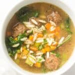 A bowl of Italian Wedding Soup with meatballs, orzo pasta, kale, diced carrots, and celery in a clear broth on a white background.