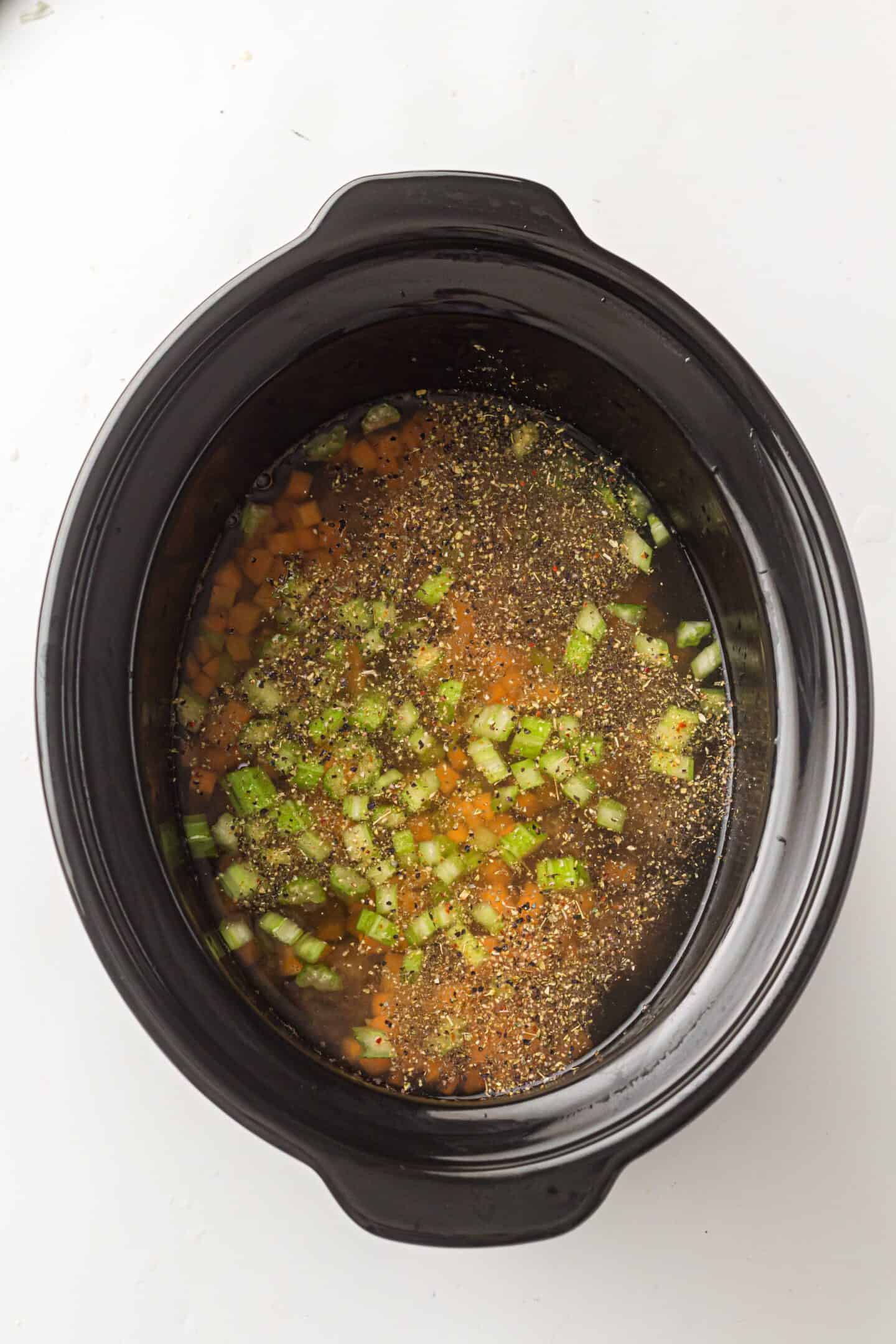 A black slow cooker filled with chopped carrots, celery, and broth makes this one-pot Italian Wedding Soup easy to prepare, with visible herbs and spices sprinkled on top.