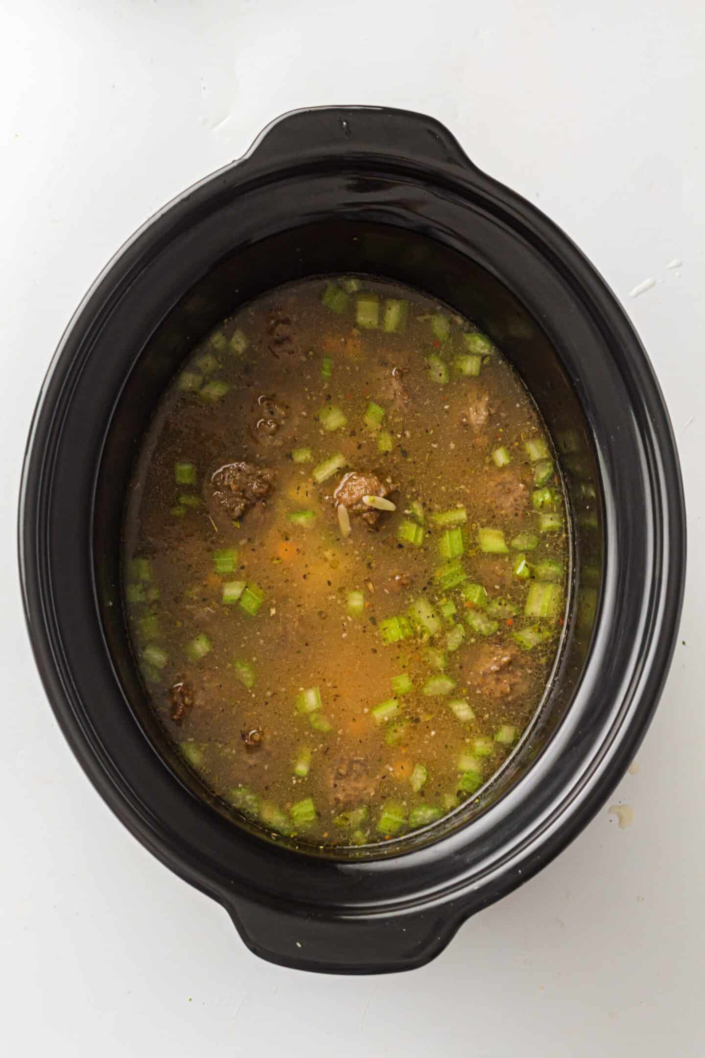 A slow cooker filled with savory beef broth, chunks of meat, and chopped celery creates an easy one-pot Italian Wedding Soup on a white background.