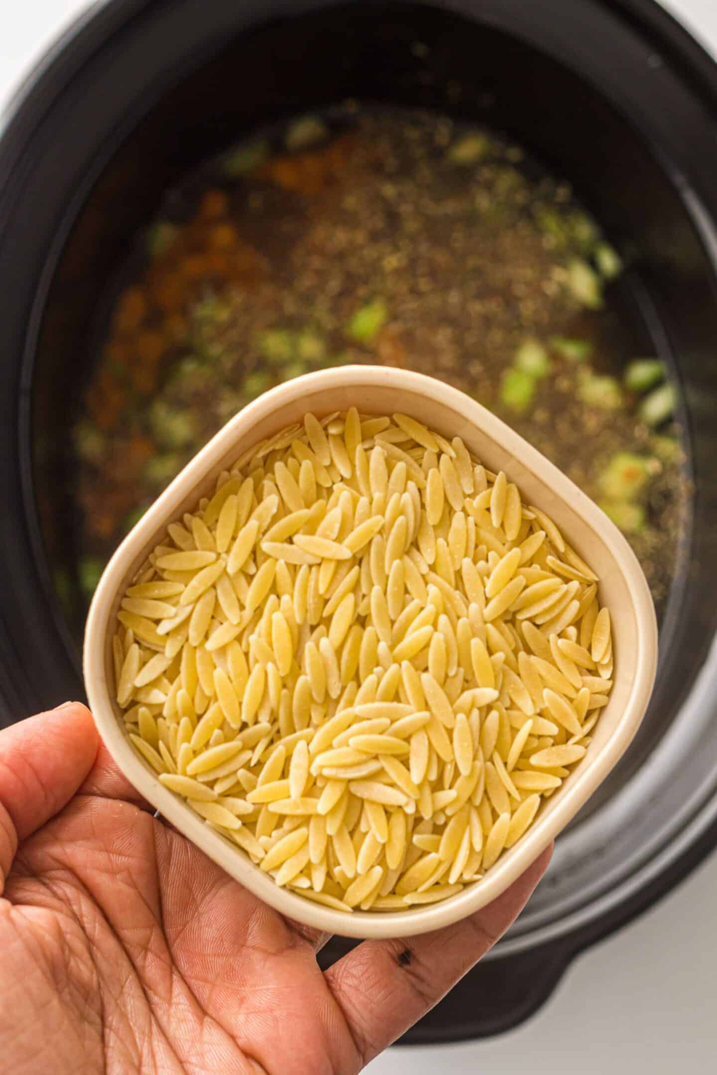 A hand holds a bowl of dry orzo pasta above a slow cooker filled with broth, seasonings, and chopped vegetables—perfect for an easy One-Pot Italian Wedding Soup.
