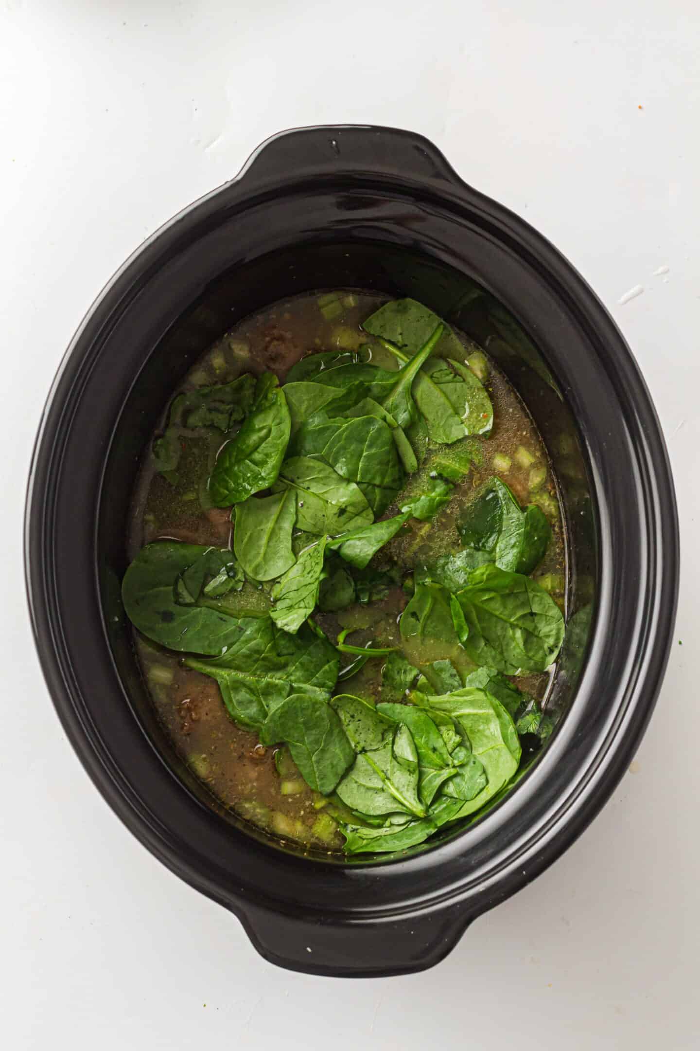 A slow cooker filled with Italian Wedding Soup ingredients, including fresh spinach leaves floating on top of a flavorful broth.