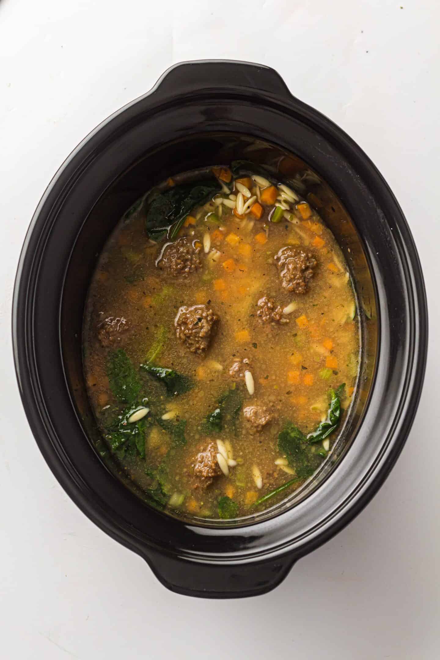 A slow cooker filled with Italian Wedding Soup featuring meatballs, chopped carrots, leafy greens, orzo pasta, and savory broth for a perfect one-pot meal.