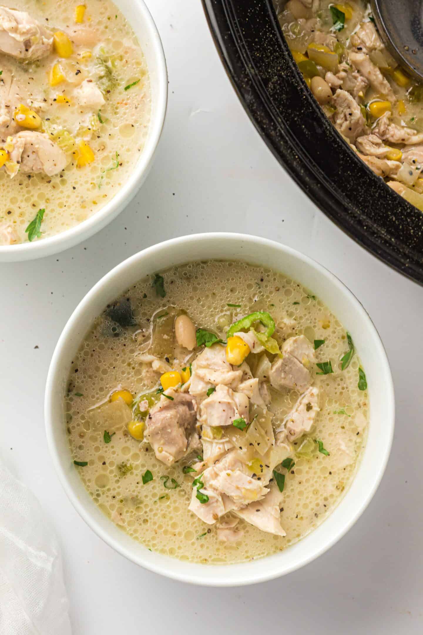 A bowl of creamy White Chicken Chili with pieces of chicken, corn, white beans, and herbs sits beside a slow cooker and another bowl filled with this hearty dish.