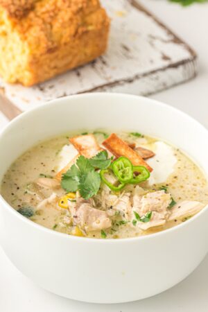 A bowl of creamy White Chicken Chili garnished with cilantro, sliced green chili, and tortilla strips, with a loaf of bread in the background.