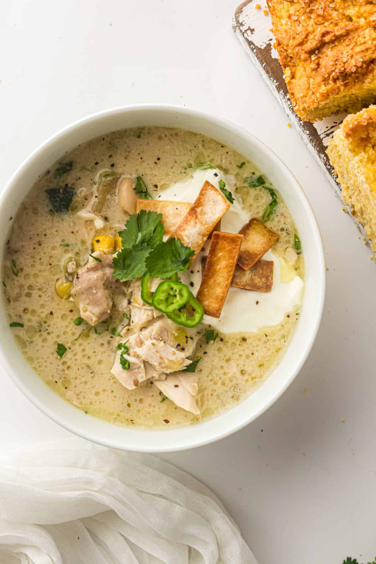 A bowl of Slow Cooker White Chicken Chili topped with sour cream, tortilla strips, cilantro, and sliced jalapeño, served next to pieces of cornbread.