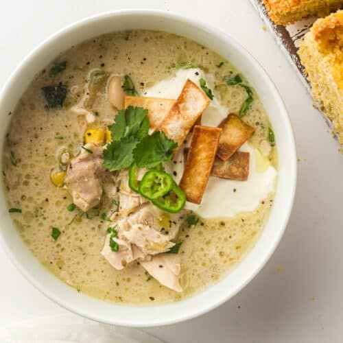 A bowl of Slow Cooker White Chicken Chili topped with sour cream, tortilla strips, cilantro, and sliced jalapeño, served next to pieces of cornbread.