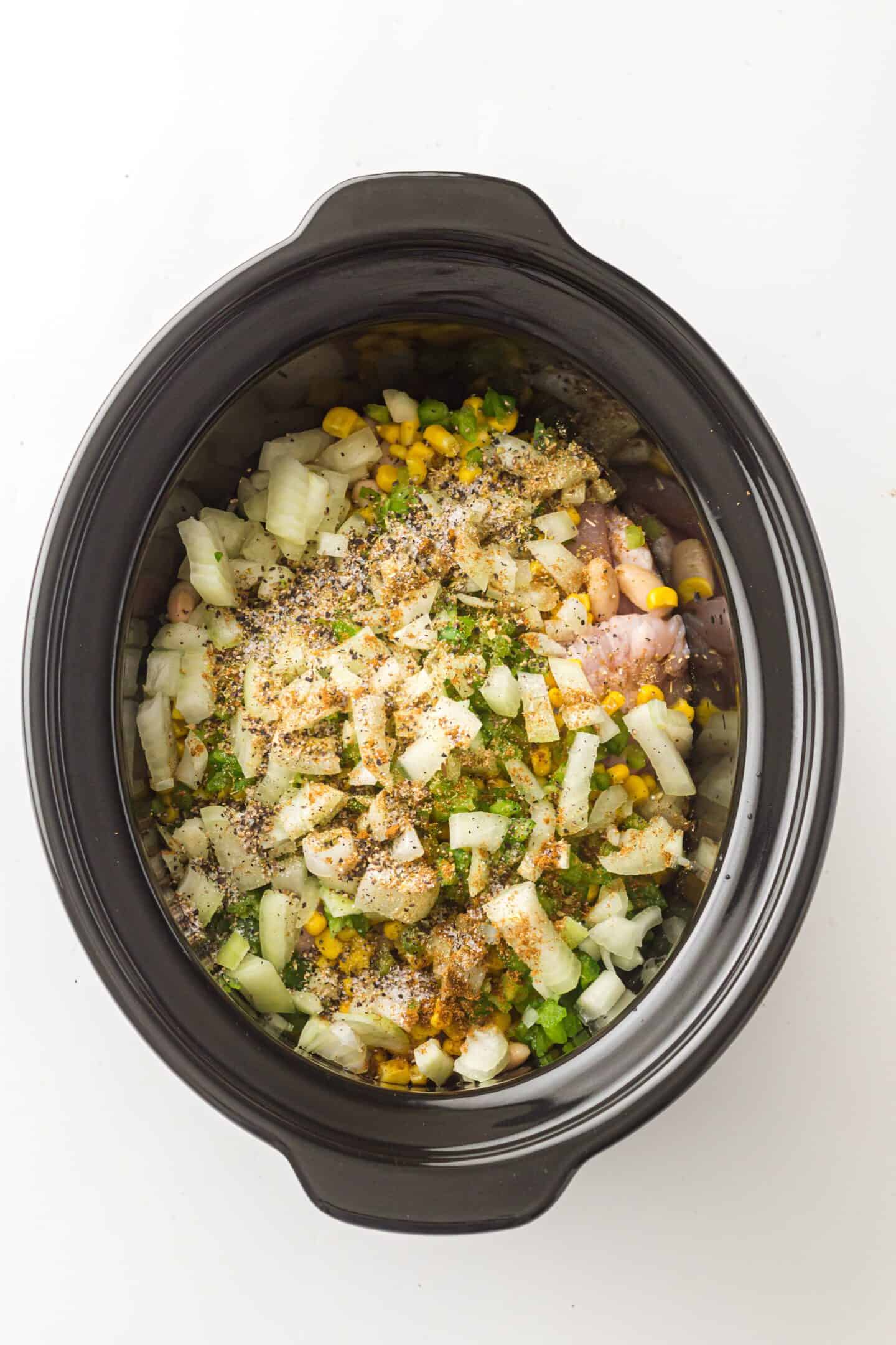 A slow cooker filled with raw ingredients, including chopped onions, corn, green peppers, beans, and seasonings over chicken—perfect for making flavorful Chicken Chili.