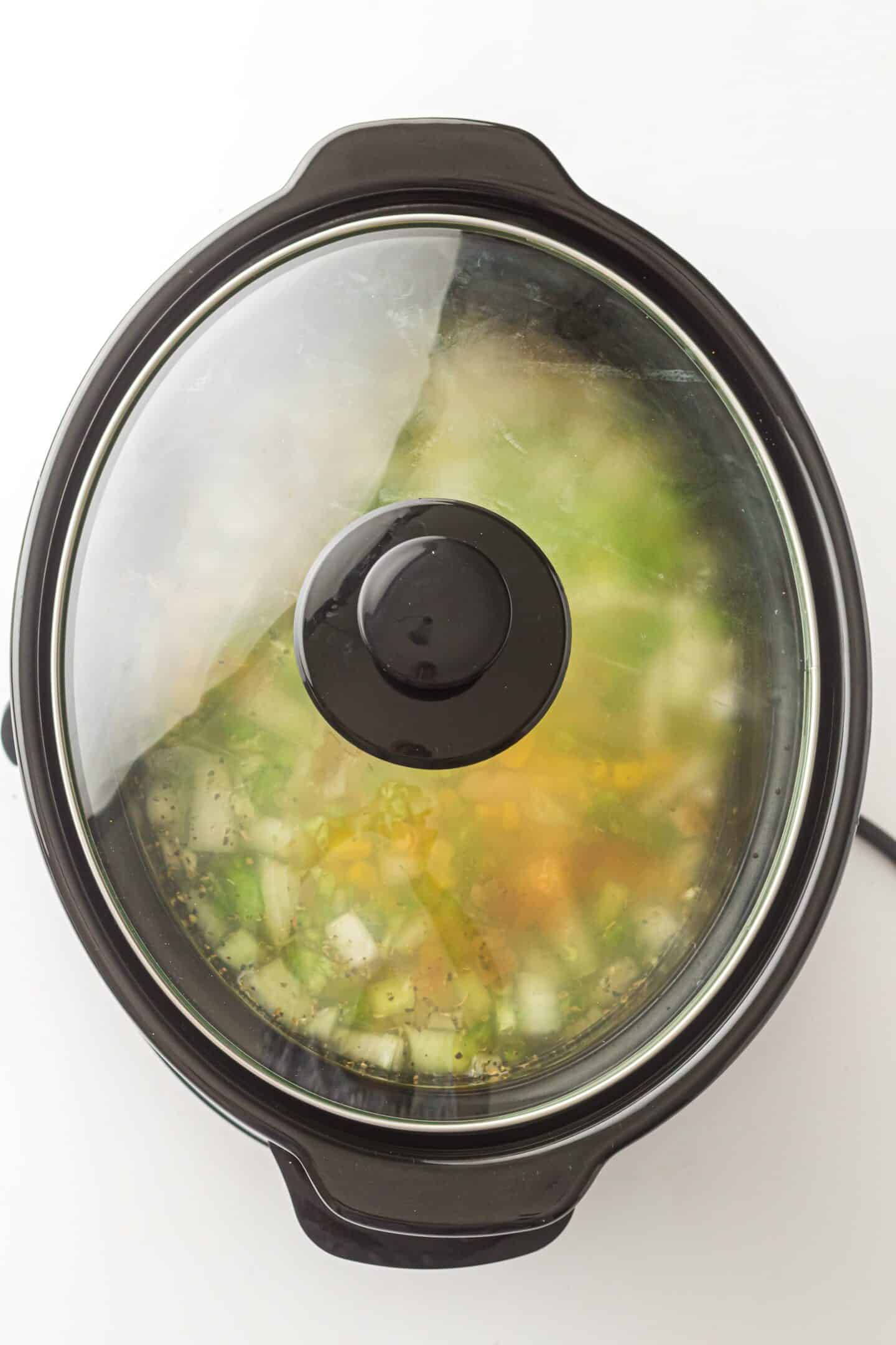 A slow cooker with a glass lid contains a hearty White Chicken Chili, featuring chopped vegetables and broth, seen from above.
