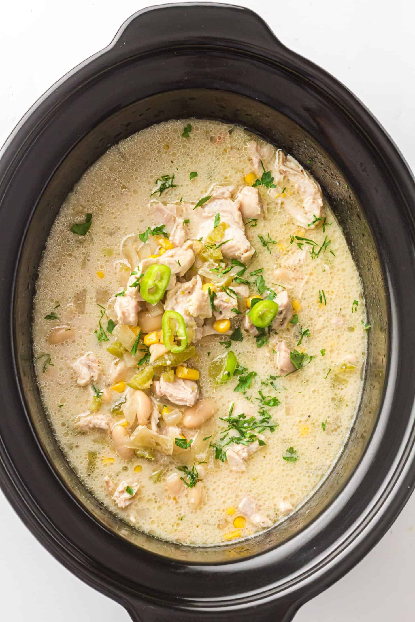A Slow Cooker brimming with creamy White Chicken Chili, packed with chunks of chicken, white beans, corn, green chilies, and finished with sliced peppers and chopped herbs.