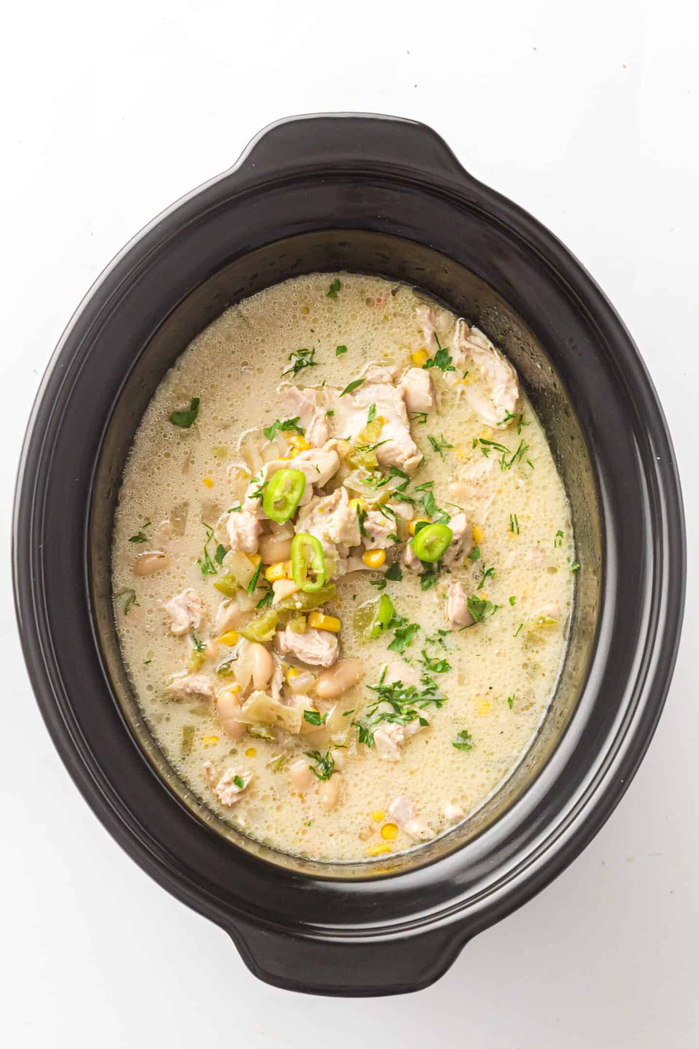 A slow cooker filled with creamy White Chicken Chili, garnished with chopped herbs and sliced green peppers.