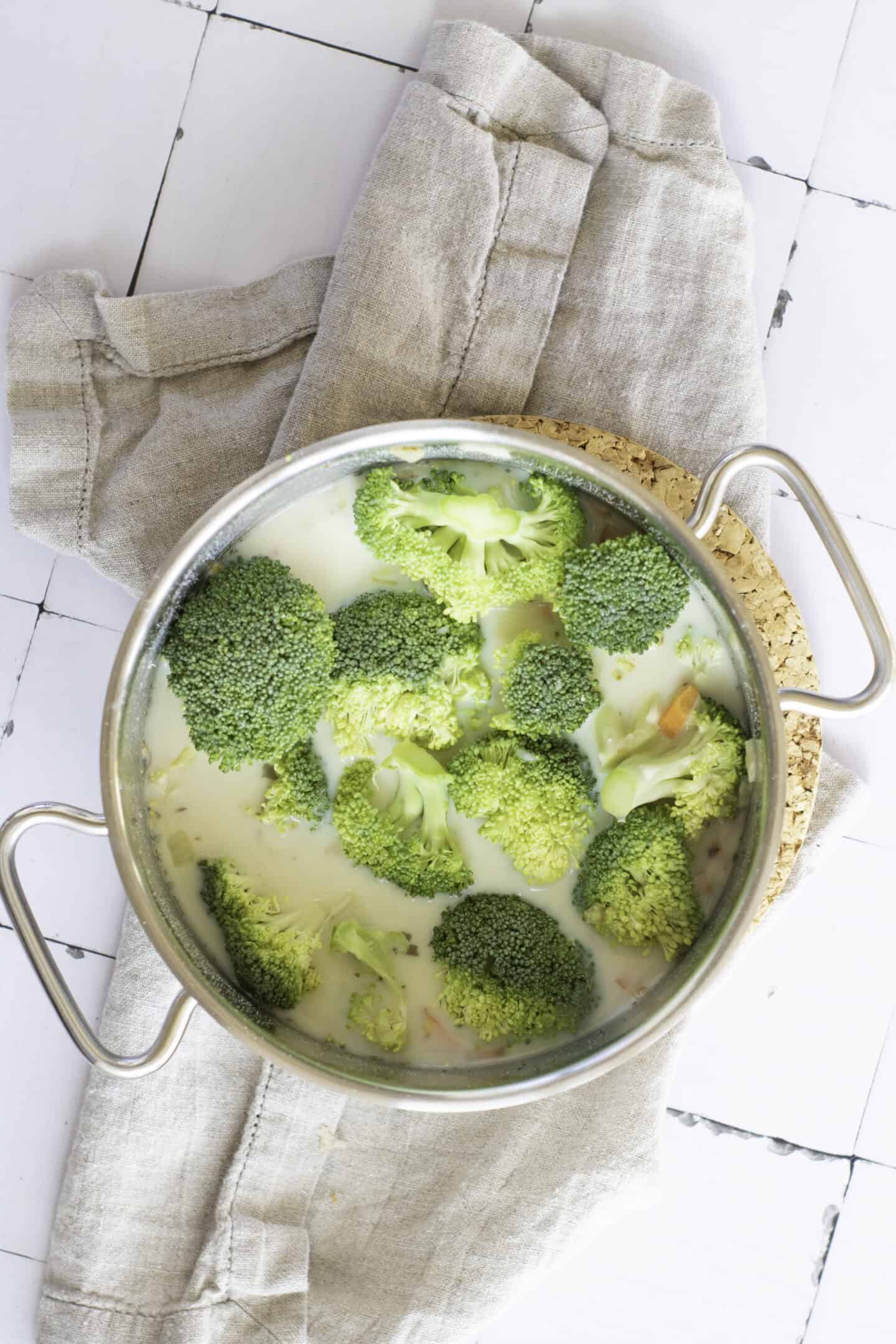 A metal pot filled with creamy Broccoli Cheddar Soup and broccoli florets sits on a beige cloth and round cork trivet, ready to be ladled into a warm sourdough bread bowl on a white tiled surface.