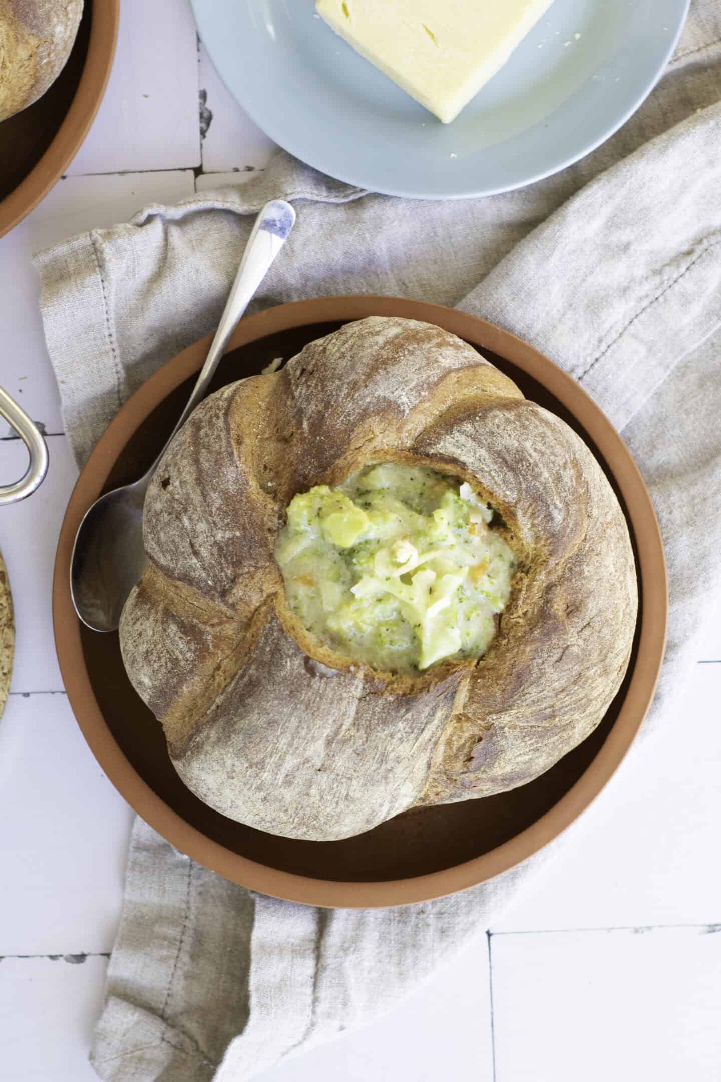 A sourdough bread bowl filled with creamy broccoli cheddar soup, placed on a terracotta plate with a spoon and a linen napkin nearby.