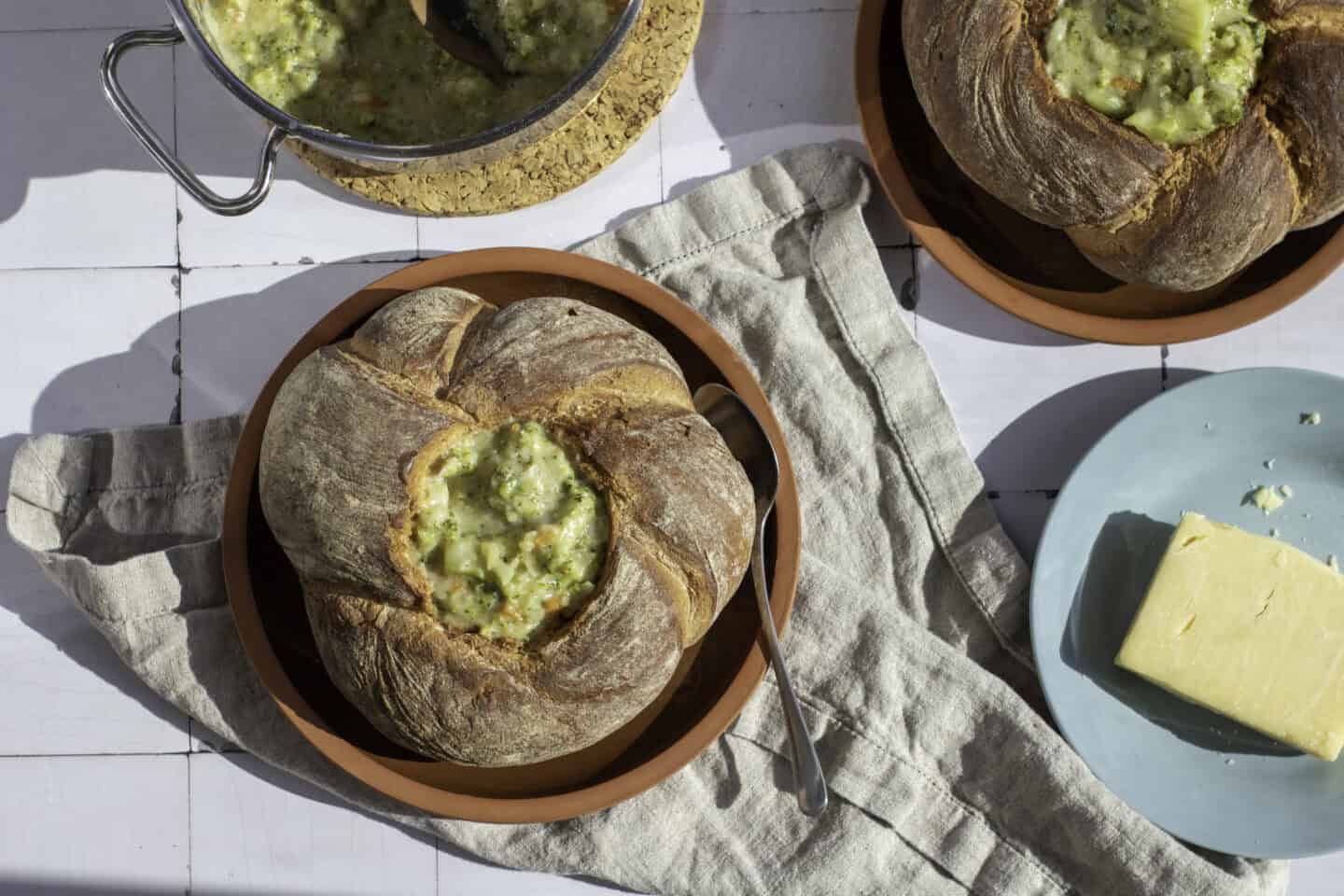 A Sourdough Bread Bowl filled with Broccoli Cheddar Soup sits on a plate with a spoon; a block of cheese and a pot of soup are also visible on the table.
