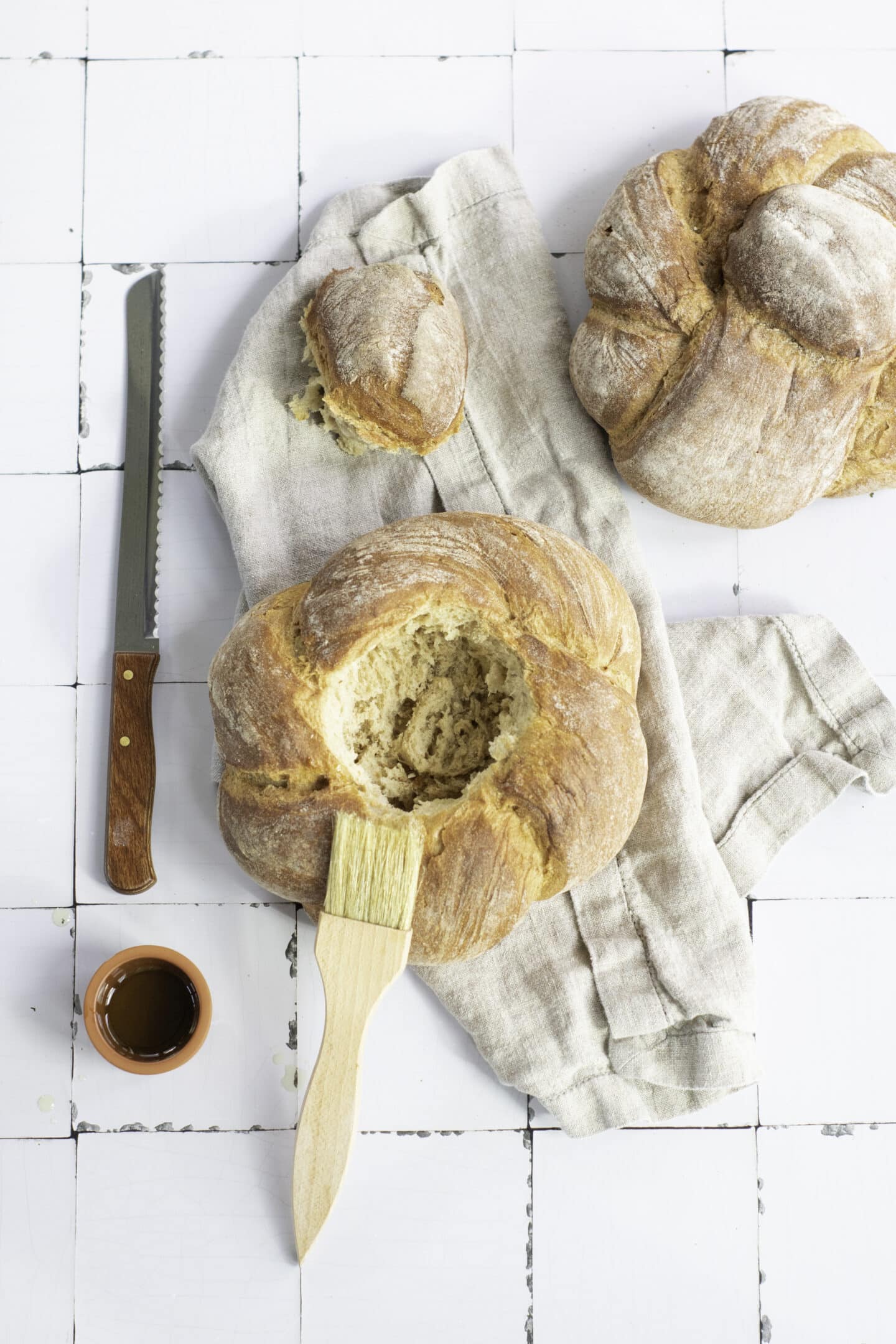 Two round loaves of rustic sourdough bread, one partially hollowed as a bread bowl, rest on a cloth. A bread knife, pastry brush, and small cup of liquid await the addition of warm broccoli cheddar soup on a white tiled surface.