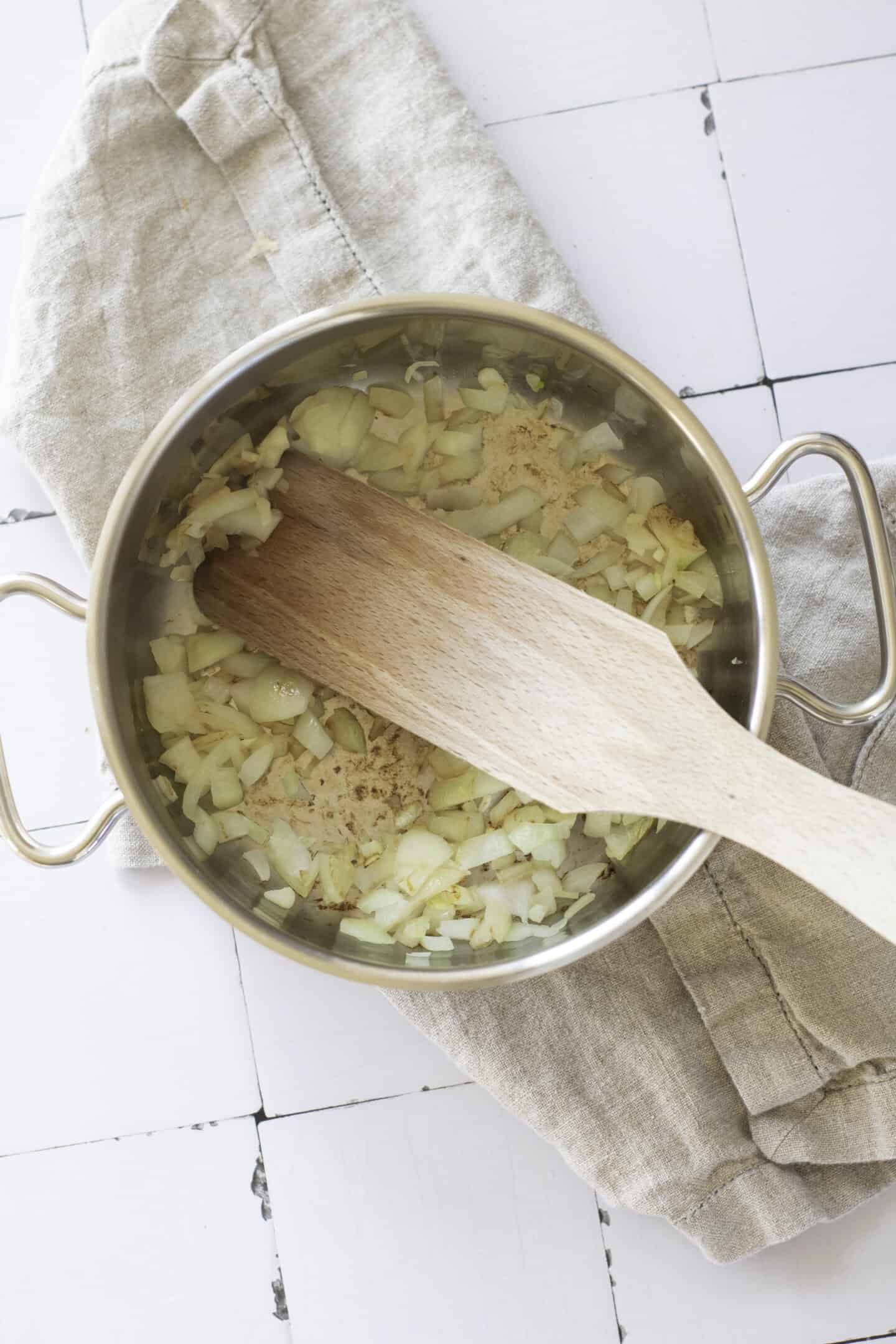 Chopped onions being sautéed in a stainless steel pot with a wooden spatula, the first step in making a creamy Broccoli Cheddar Soup, placed on a beige cloth on a white tiled surface.