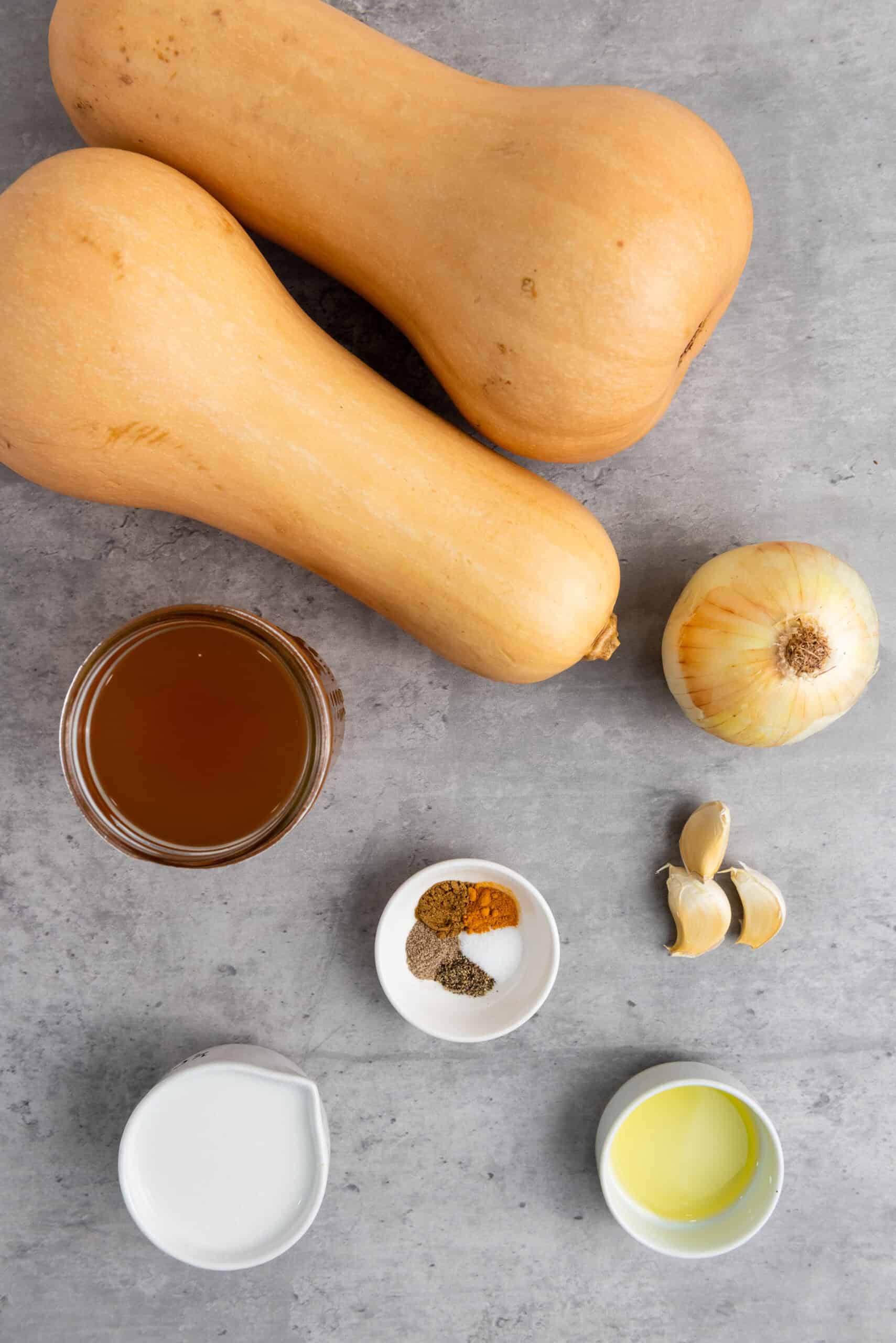 For a cozy Butternut Squash Soup, two whole butternut squashes, a yellow onion, garlic cloves, broth, spices, oil, and cream are arranged on a gray countertop—perfect ingredients for your slow cooker.
