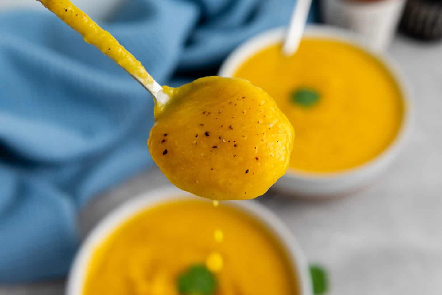 A close-up of a spoonful of butternut squash soup with visible black pepper, held above a bowl of the same soup with a garnish, and a blue cloth in the background—perfect for cozy slow cooker meals.