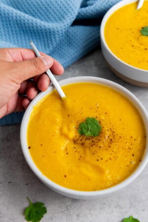 A hand holds a spoon in a bowl of creamy yellow Butternut Squash Soup, garnished with black pepper and a sprig of cilantro, with another bowl visible in the background.