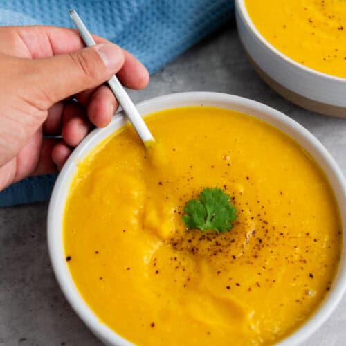 A hand holds a spoon in a bowl of creamy yellow Butternut Squash Soup, garnished with black pepper and a sprig of cilantro, with another bowl visible in the background.