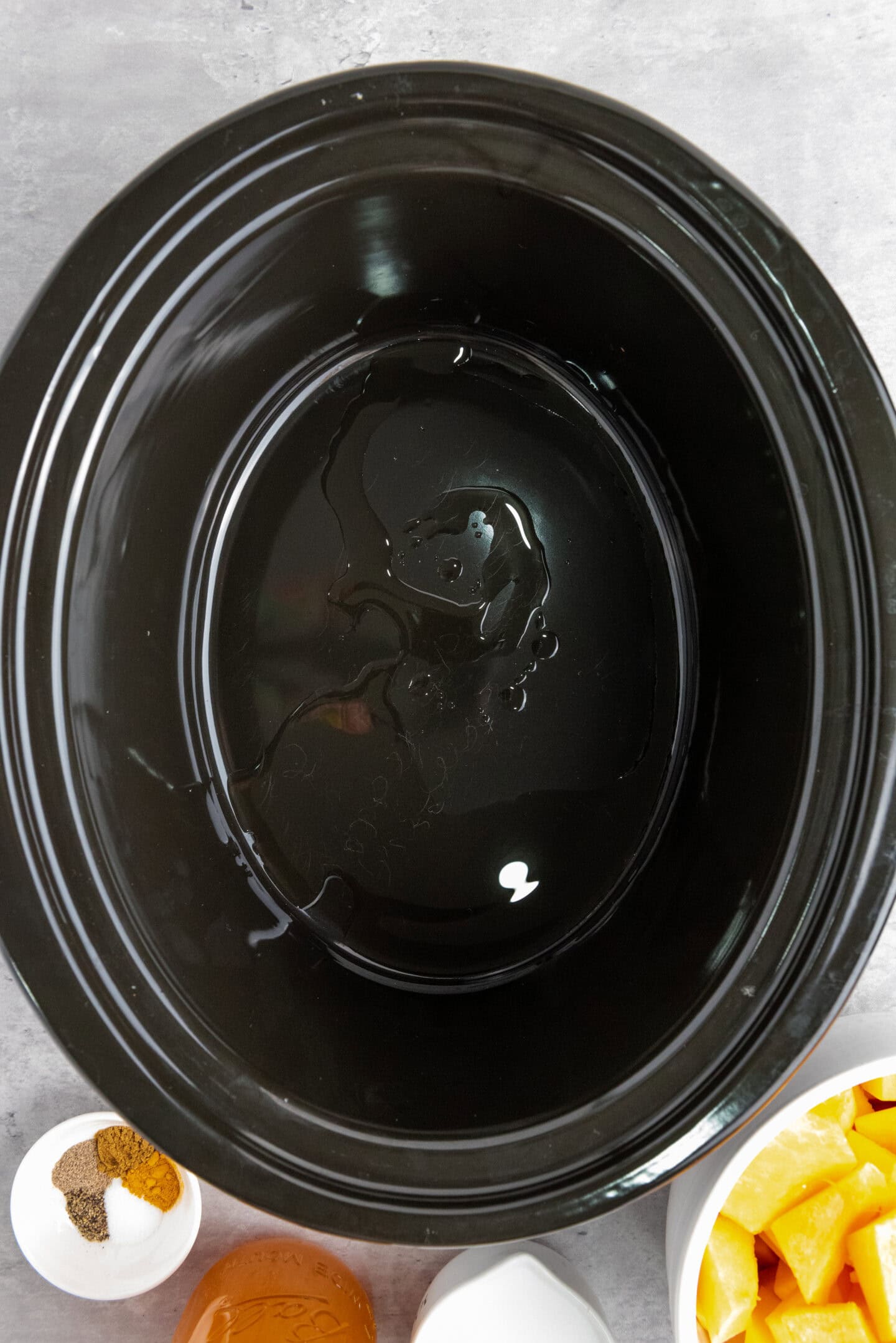 A black slow cooker with a small amount of oil inside, surrounded by small bowls containing spices, honey, and chopped vegetables—perfect for preparing a flavorful butternut squash soup.