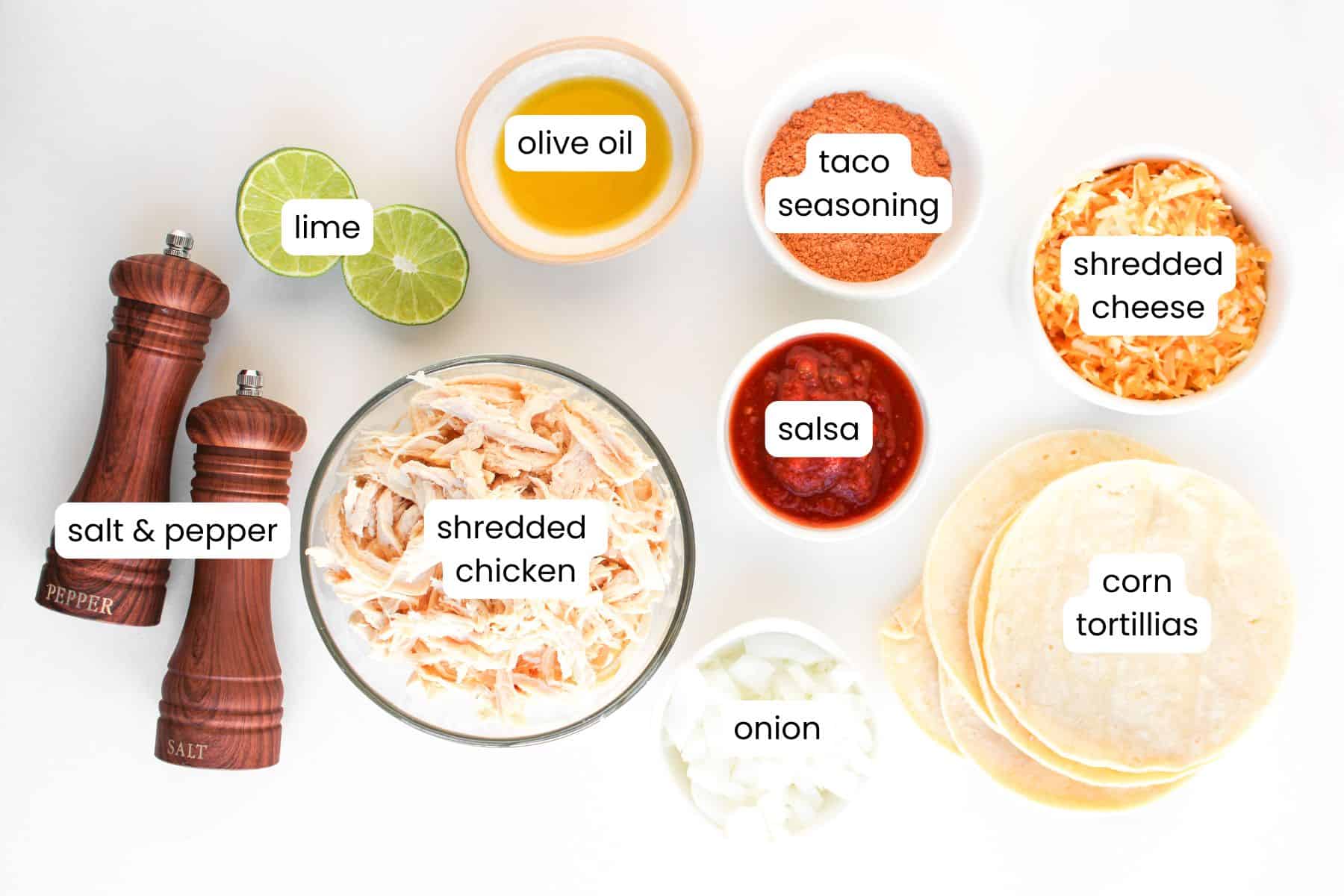 Flat lay of labeled taco ingredients—perfect for Crispy Baked Chicken Tacos—including salt, pepper, lime, olive oil, taco seasoning, shredded cheese, shredded chicken, salsa, chopped onion, and corn tortillas on a white background.