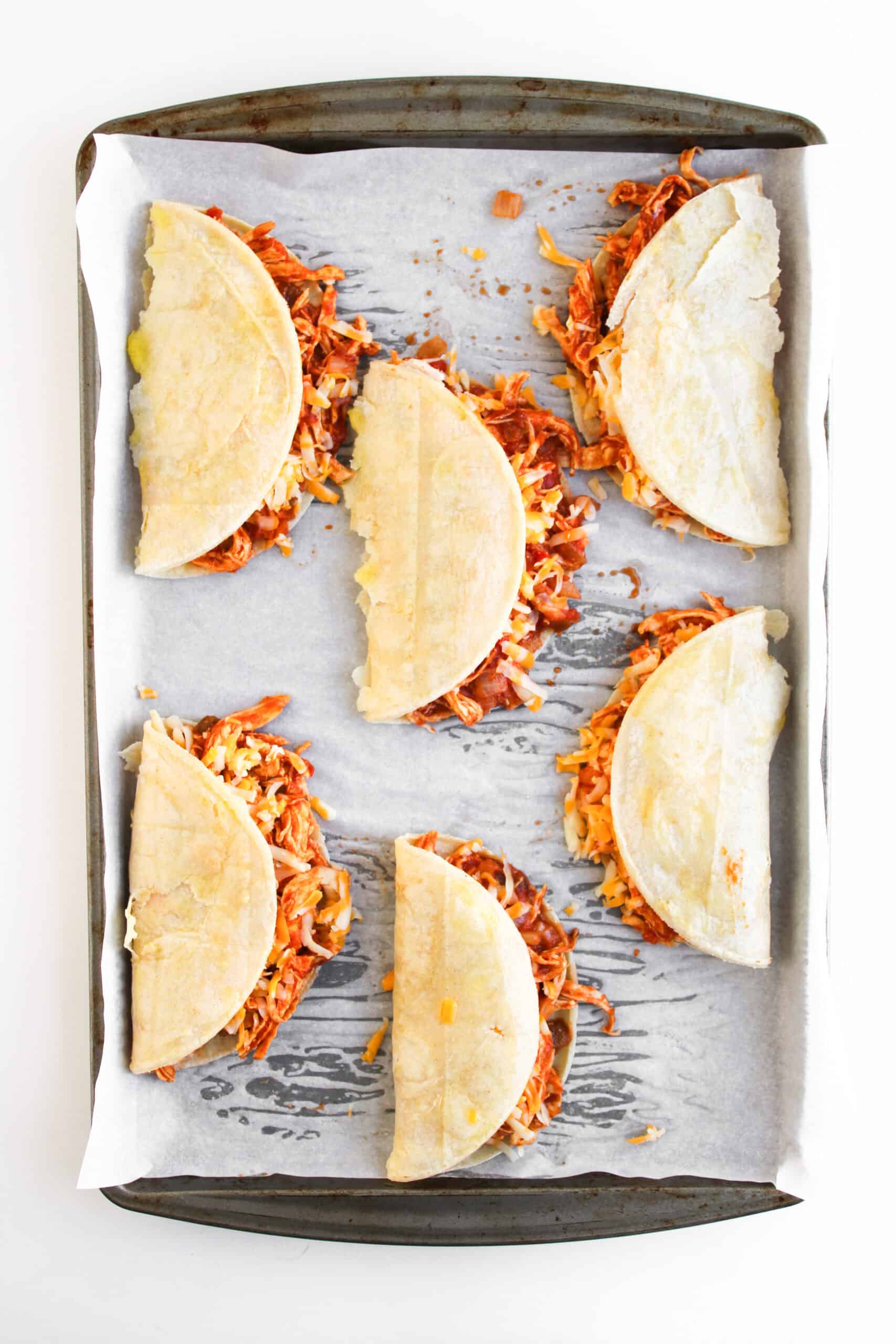Six folded tortillas filled with shredded, cooked meat and cheese are arranged on a parchment-lined baking sheet—perfect for making Crispy Baked Chicken Tacos.