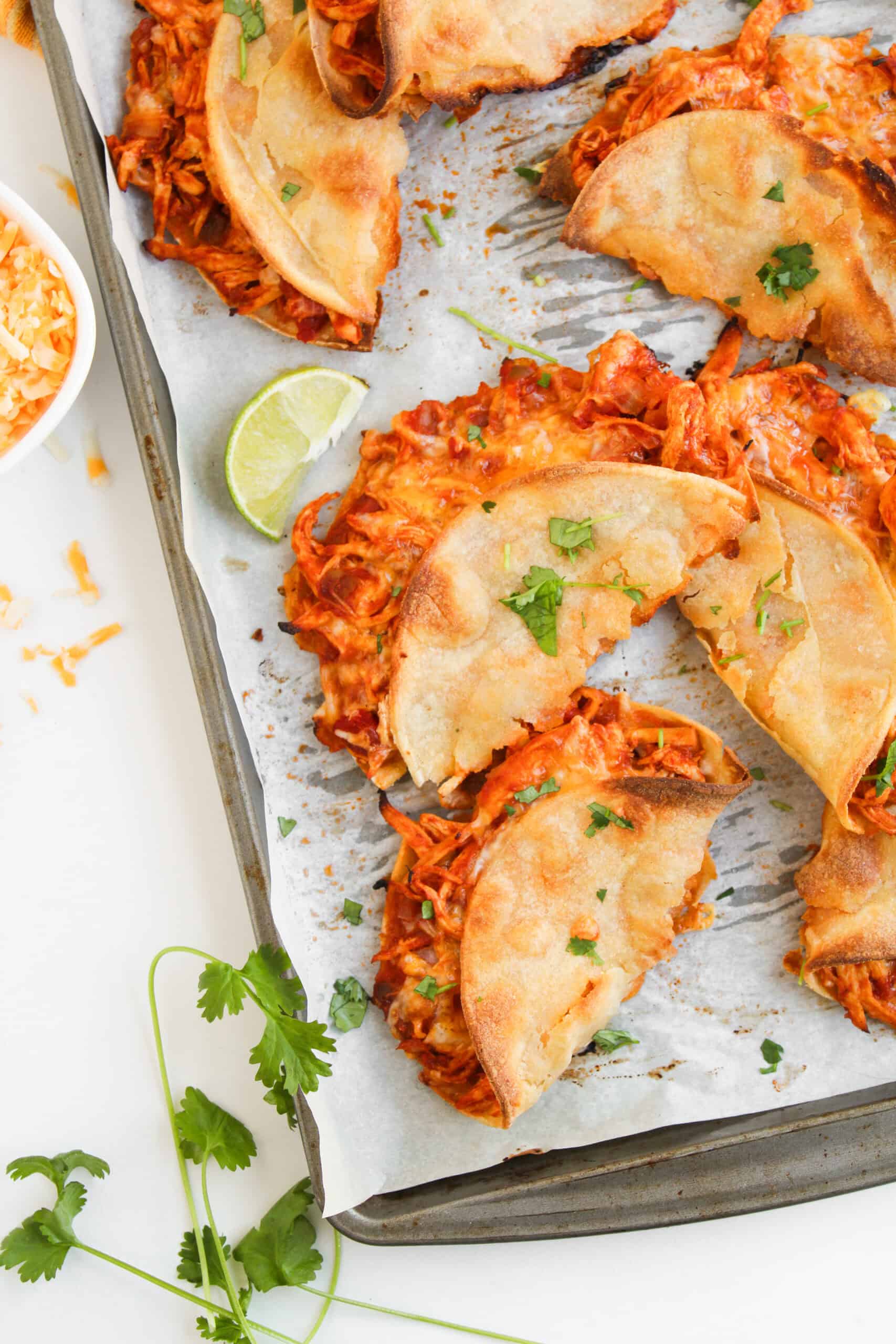 Crispy Baked Chicken Tacos arranged on a parchment-lined baking sheet, garnished with chopped cilantro, served with a lime wedge and shredded cheese on the side.