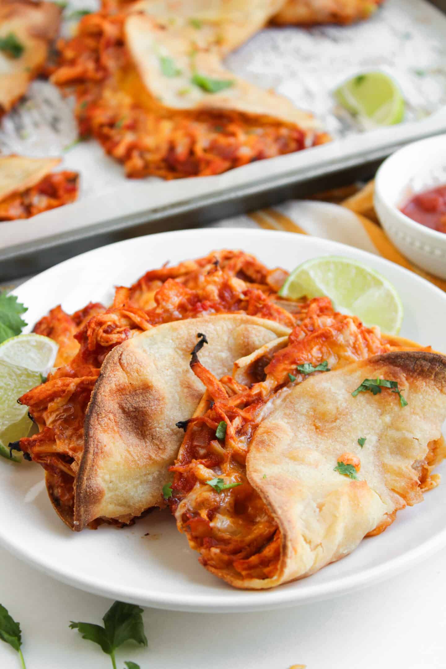 Two baked chicken tacos on a white plate, garnished with chopped herbs and lime wedges, with more crispy chicken tacos and a bowl of salsa in the background.