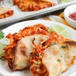 Two baked chicken tacos on a white plate, garnished with chopped herbs and lime wedges, with more crispy chicken tacos and a bowl of salsa in the background.