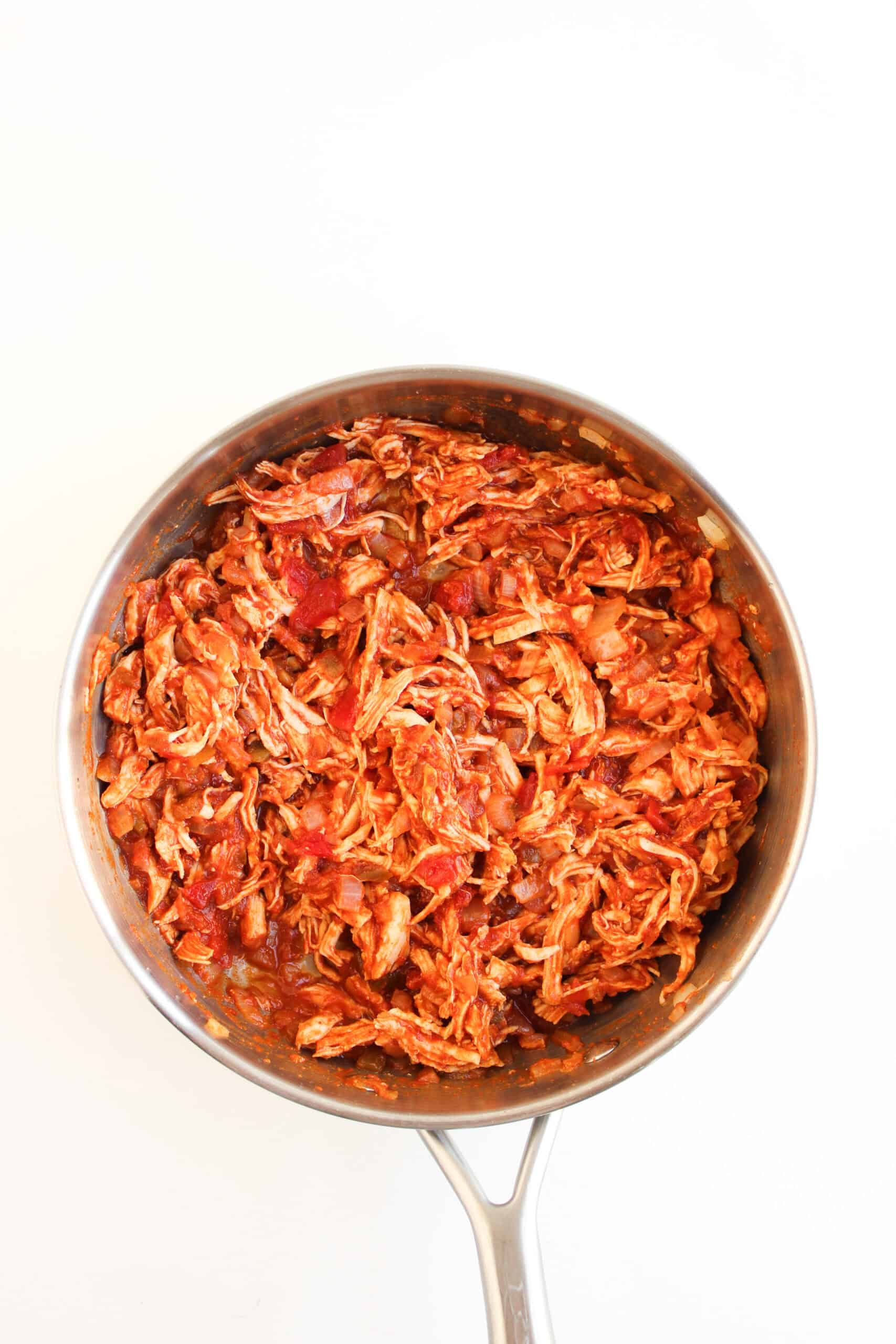 A stainless steel pan filled with shredded chicken mixed in a red tomato-based sauce, perfect for stuffing into Crispy Baked Chicken Tacos, viewed from above on a white background.