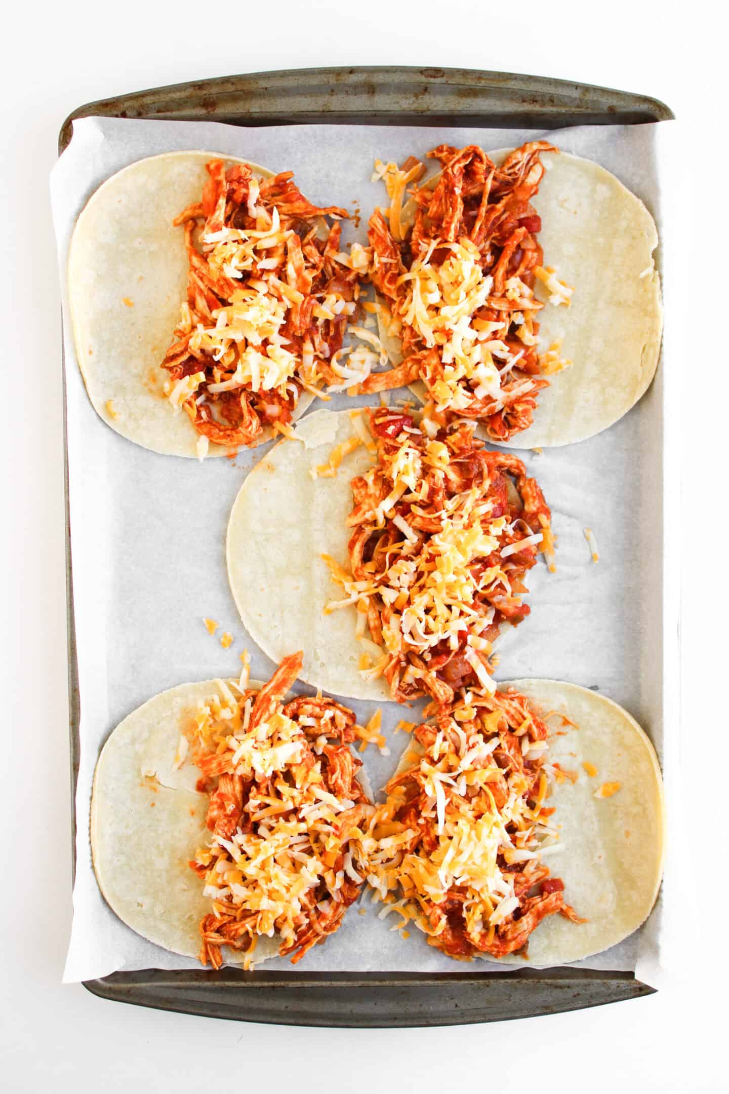 A baking sheet lined with parchment paper holds five corn tortillas topped with shredded chicken and cheese—perfect for making Crispy Baked Chicken Tacos.