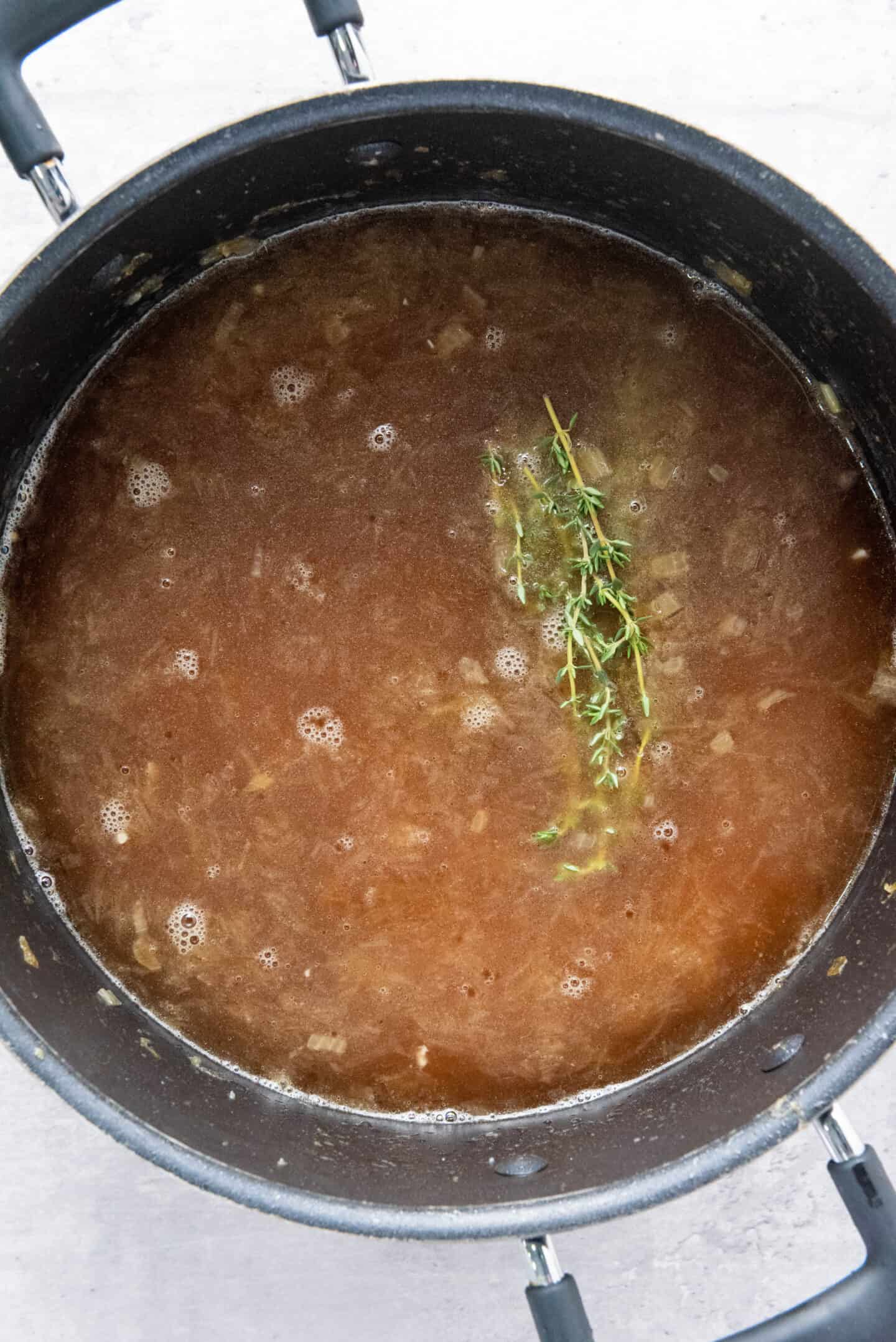 A pot of French Onion Soup, featuring brown broth with visible onions and a sprig of fresh thyme floating on top.