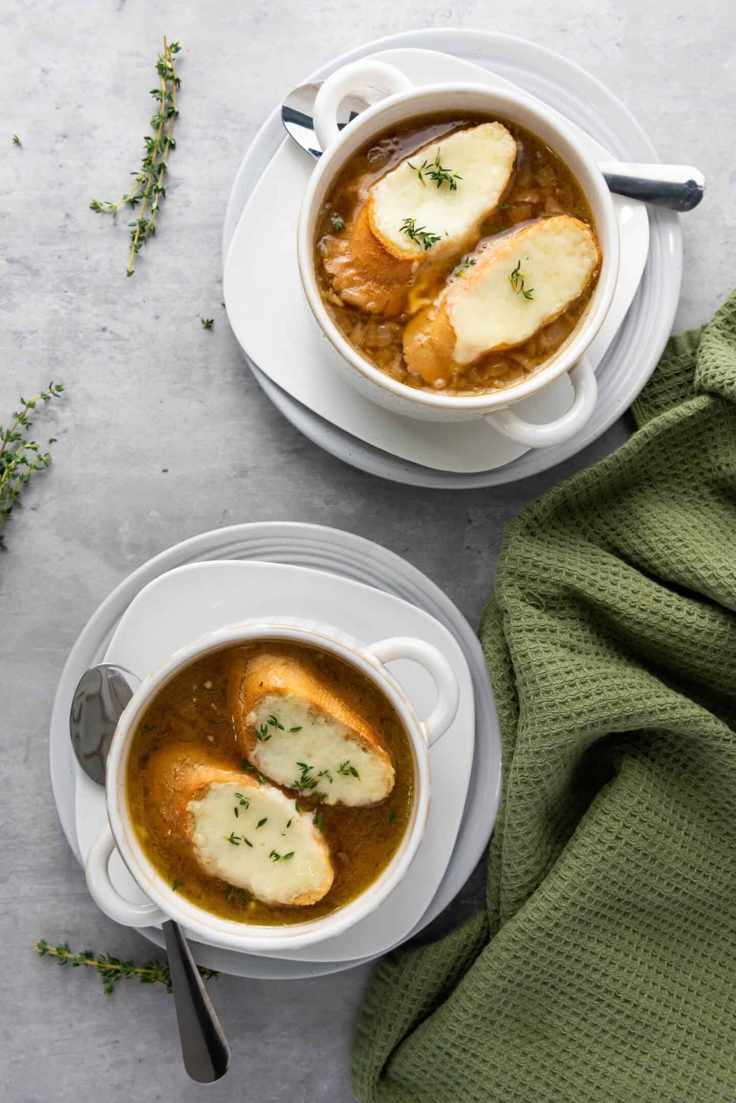 Two bowls of classic French Onion Soup topped with toasted bread and melted cheese, served on white plates with spoons; sprigs of thyme and a green cloth are nearby.
