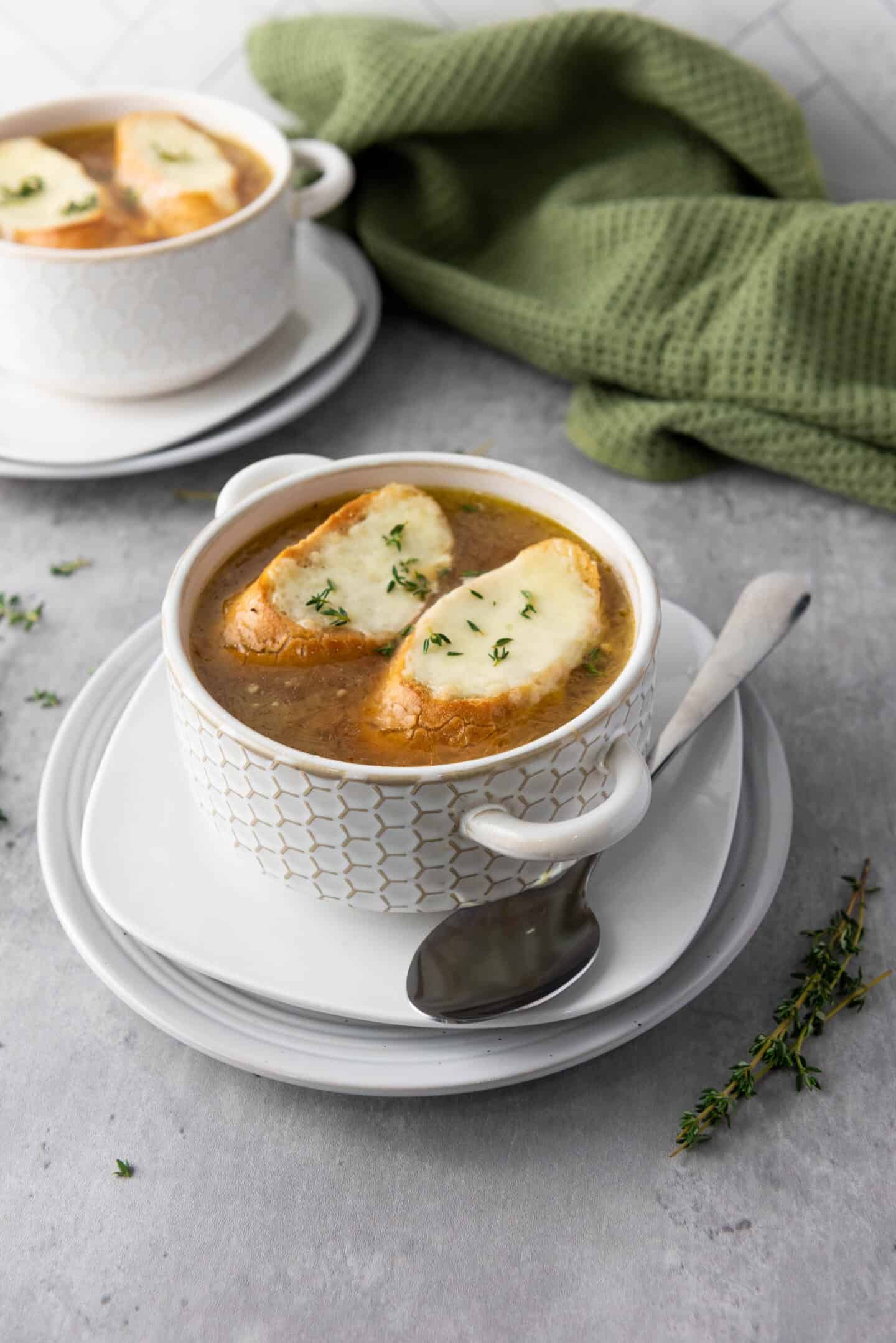 A bowl of French onion soup topped with two slices of bread and melted cheese, served on a plate with a spoon; another bowl and a green cloth are in the background.