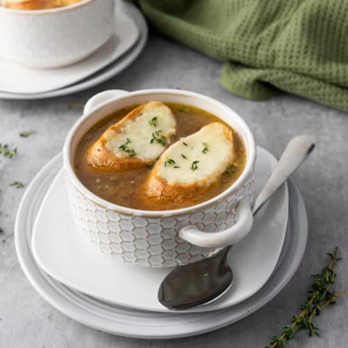A bowl of French onion soup topped with two slices of bread and melted cheese, served on a plate with a spoon; another bowl and a green cloth are in the background.