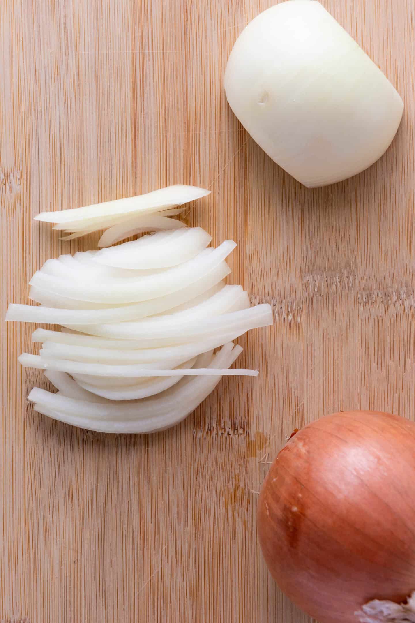 A half-peeled onion, a halved onion, and thinly sliced onion pieces are on a wooden cutting board—perfect prep for classic French Onion Soup.